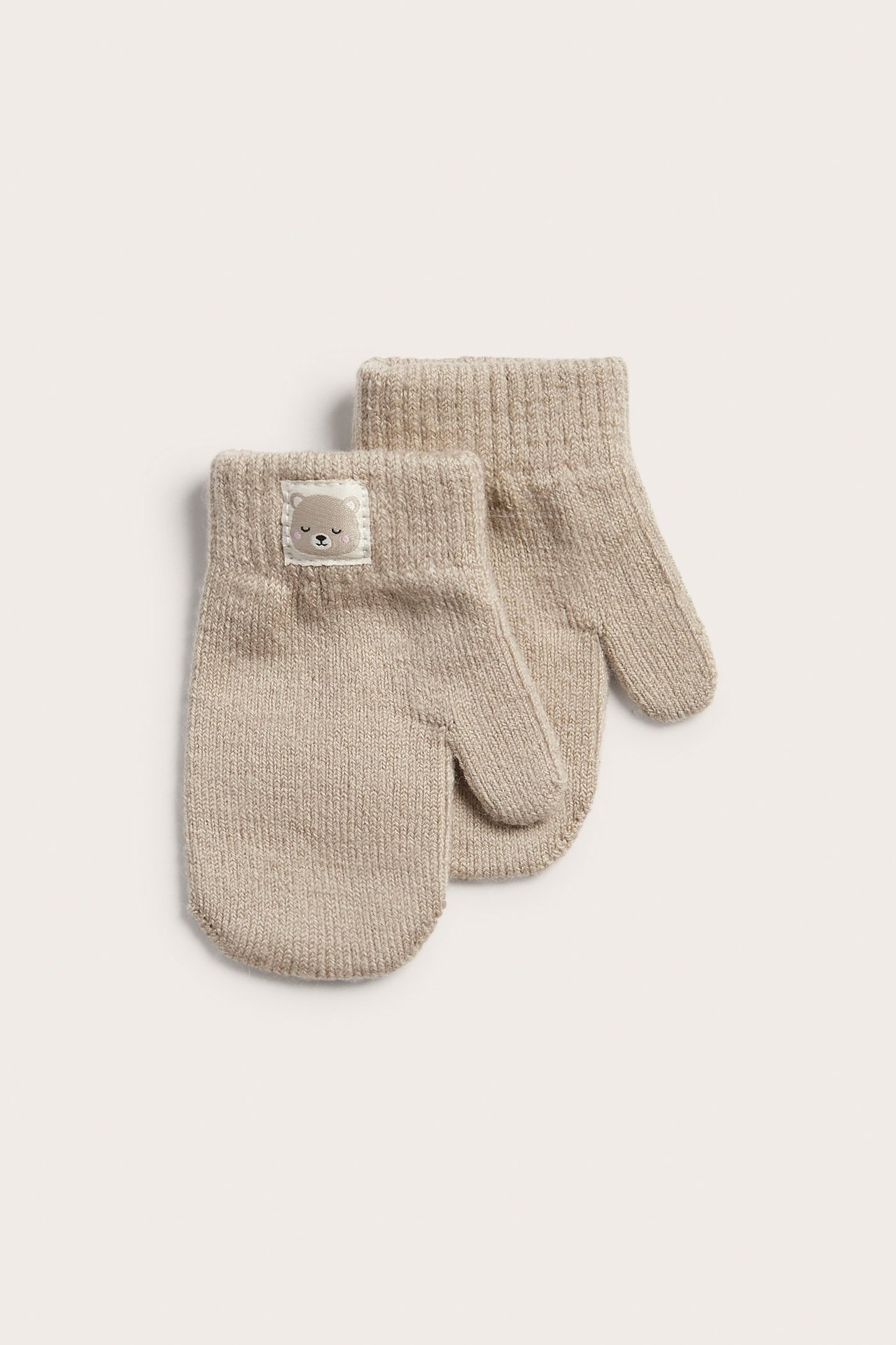 Framifrån: två beiga stickade babyvantar i ullmix med ribbade muddar och en liten björnetikett.
