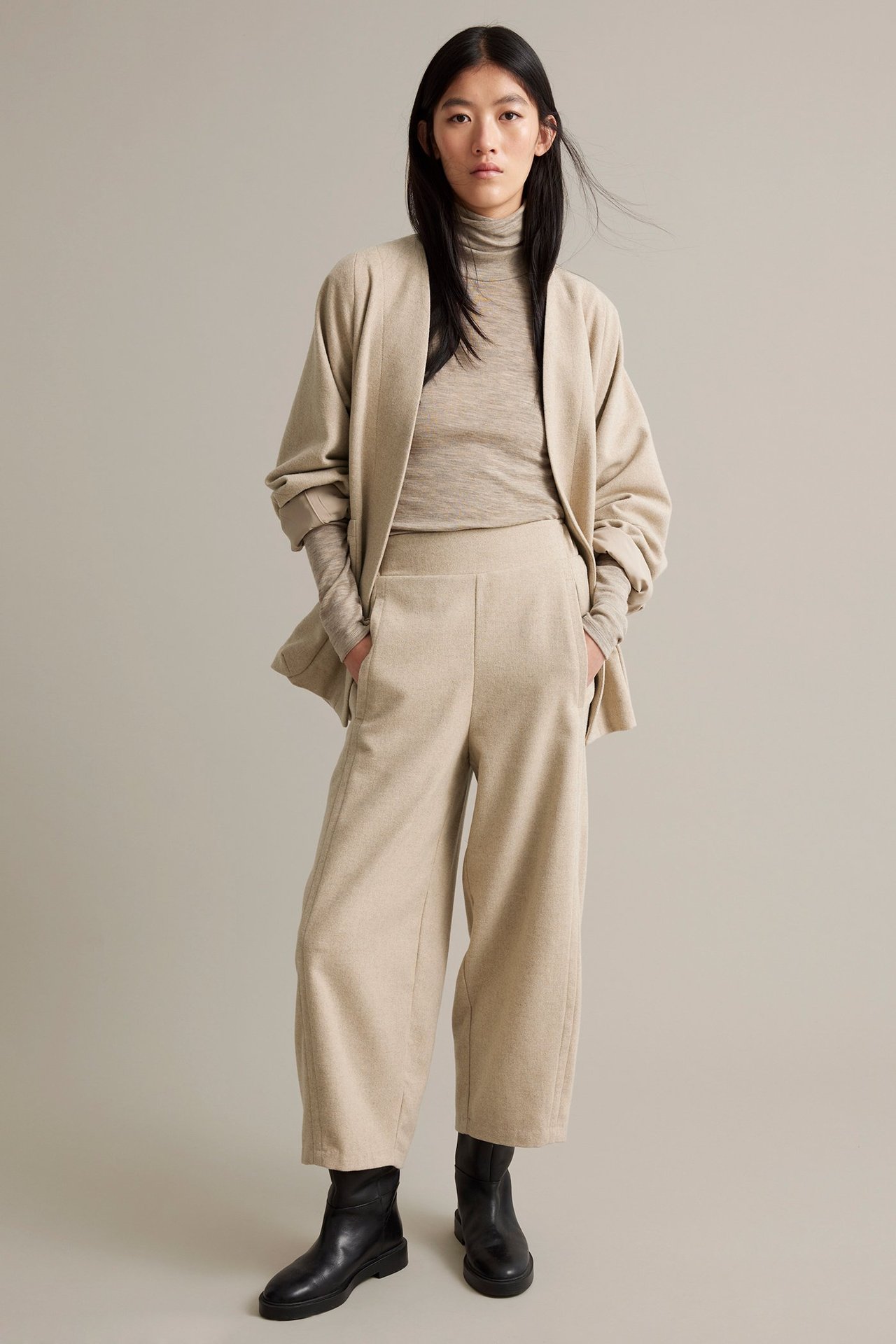 Nainen beigeä melange-villasekoitehousuissa, takataskut, elastinen vyötärö, sopiva yläosa ja mustat saappaat.