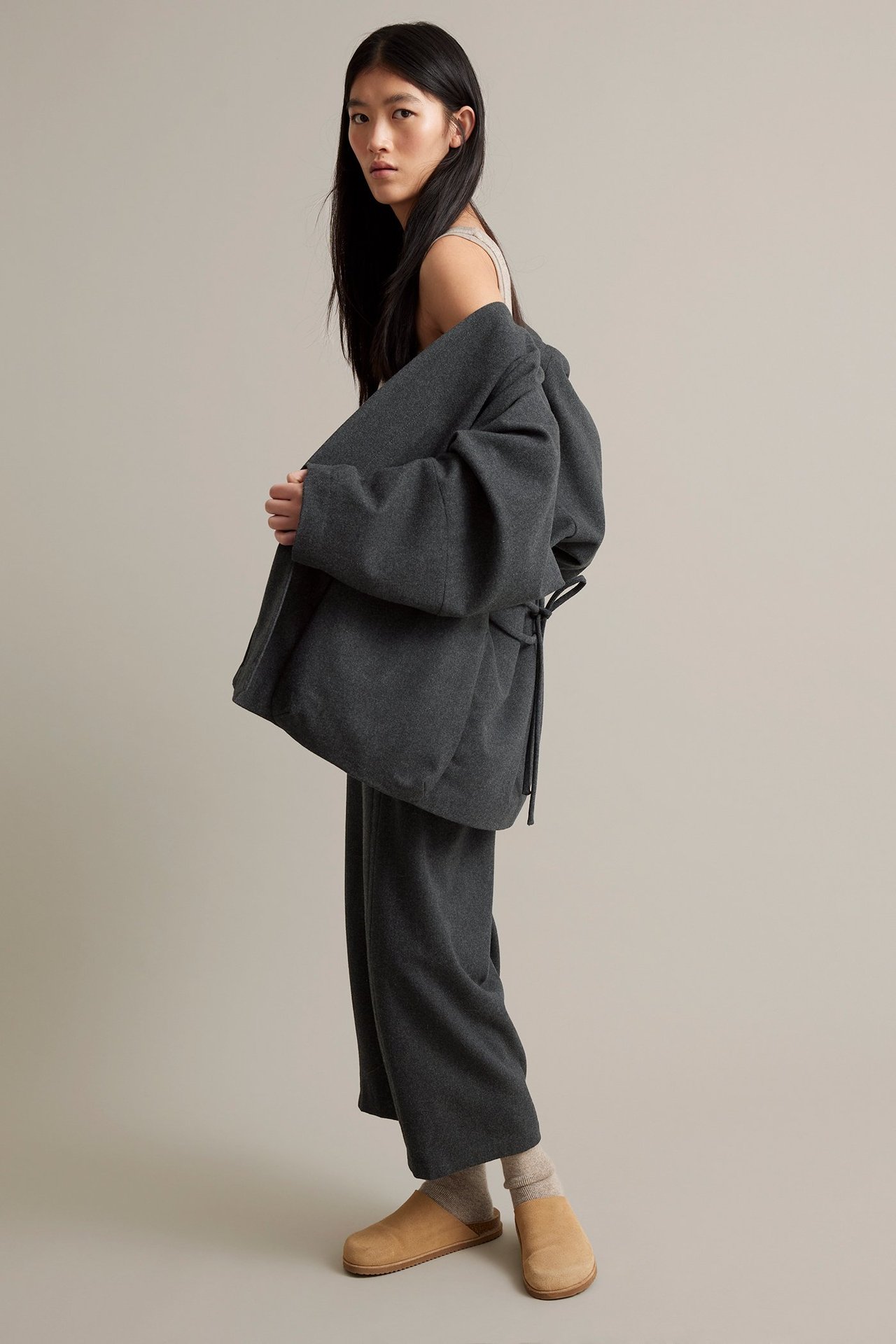 Nuori nainen tummanharmaissa villasekoitteisissa housuissa, iso harmaa takki ja beige jalkineet.