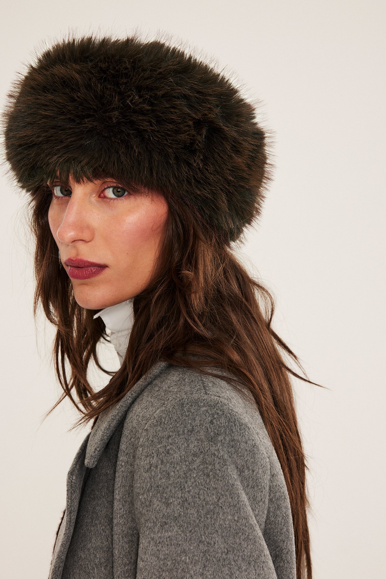 Lähikuva naisesta, jolla on ruskea tekoturkis hiuspanta, valkoinen poolopaita ja harmaa takki.