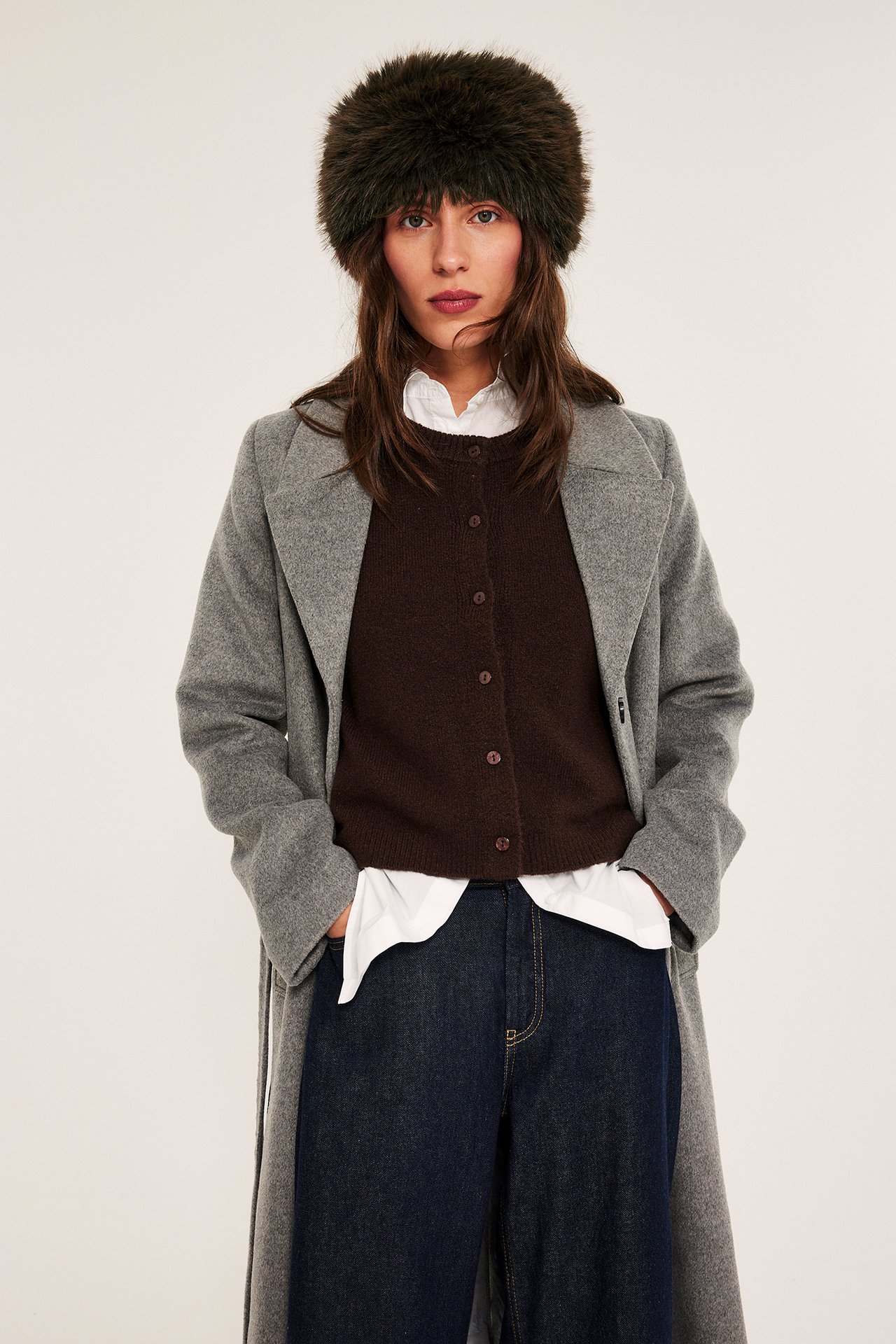 Edestä: Nainen yllään ruskea tekoturkis hiuspanta, harmaa takki, ruskea neuletakki, valkoinen paita ja farkut.