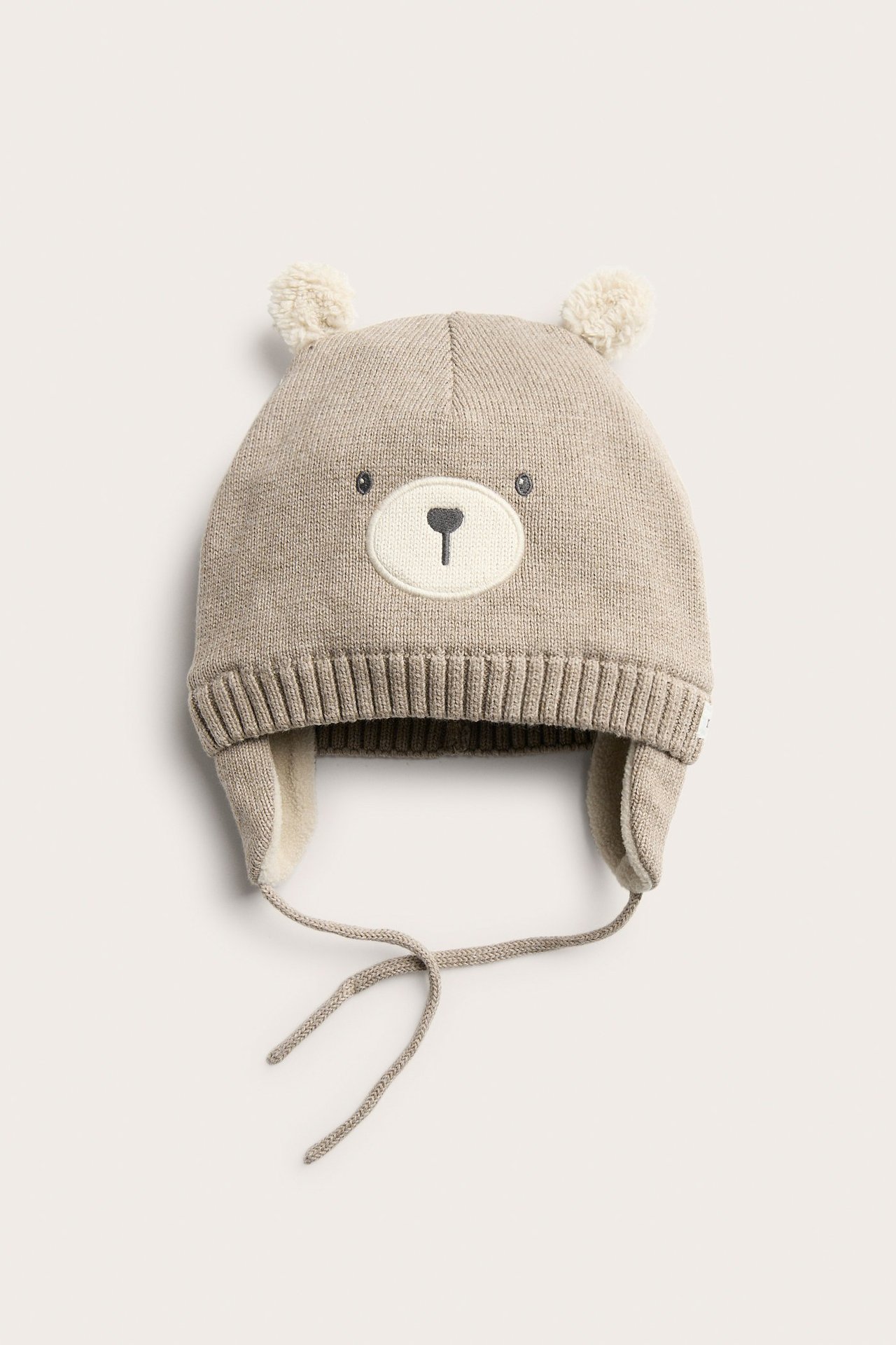 Widok z przodu beżowej melanżowej dzianinowej czapki niemowlęcej z twarzą misia, pomponami, nausznikami i wiązaniami.