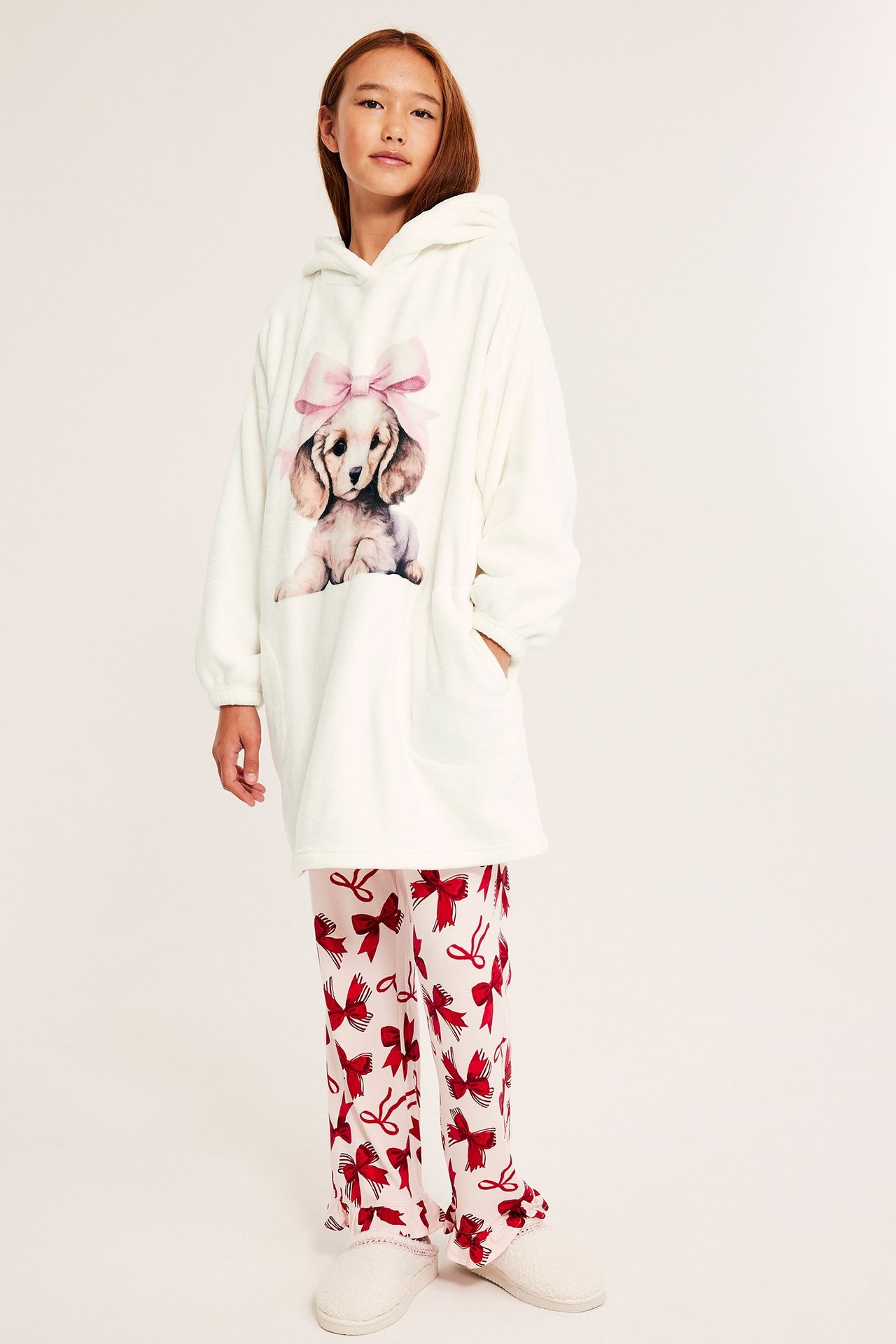 Edestäpäin: lapsi yllään luonnonvalkoinen oversized fleecehuppari koiraprintillä, punarusettiset pyjamahousut ja valkoiset tossut.