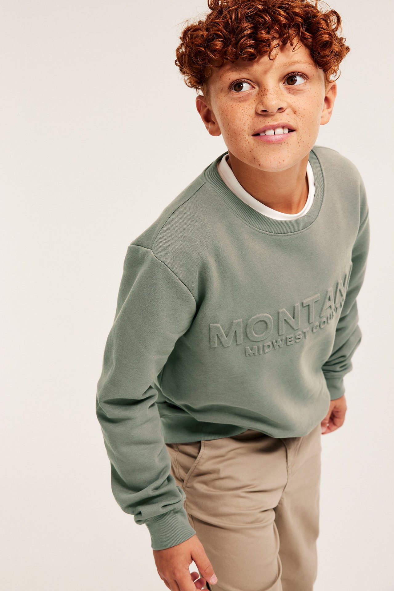 Chłopak w szarej bluzie z nadrukiem 3D, beżowe spodnie, kręcone włosy, lekki uśmiech.