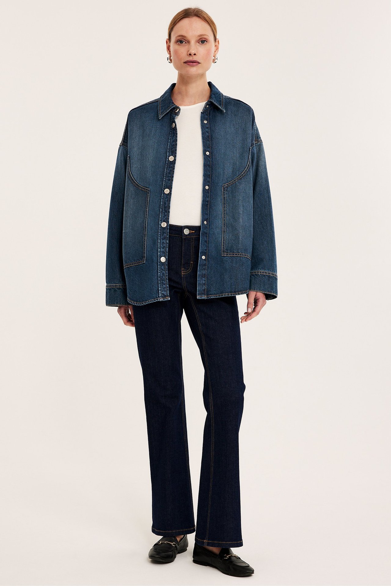 Framifrån: Kvinna bär en oversized blå jeansskjorta med tryckknappar, vit t-shirt, mörka jeans, svarta loafers.