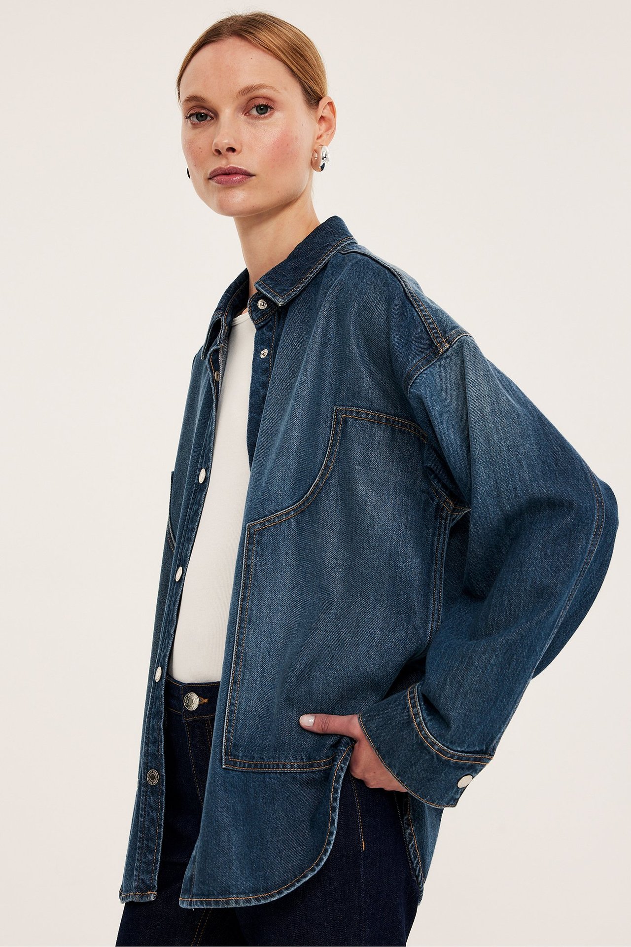 Framifrån: Kvinna i oversized blå jeansskjorta med stora fickor, tryckknappar, över vit t-shirt.