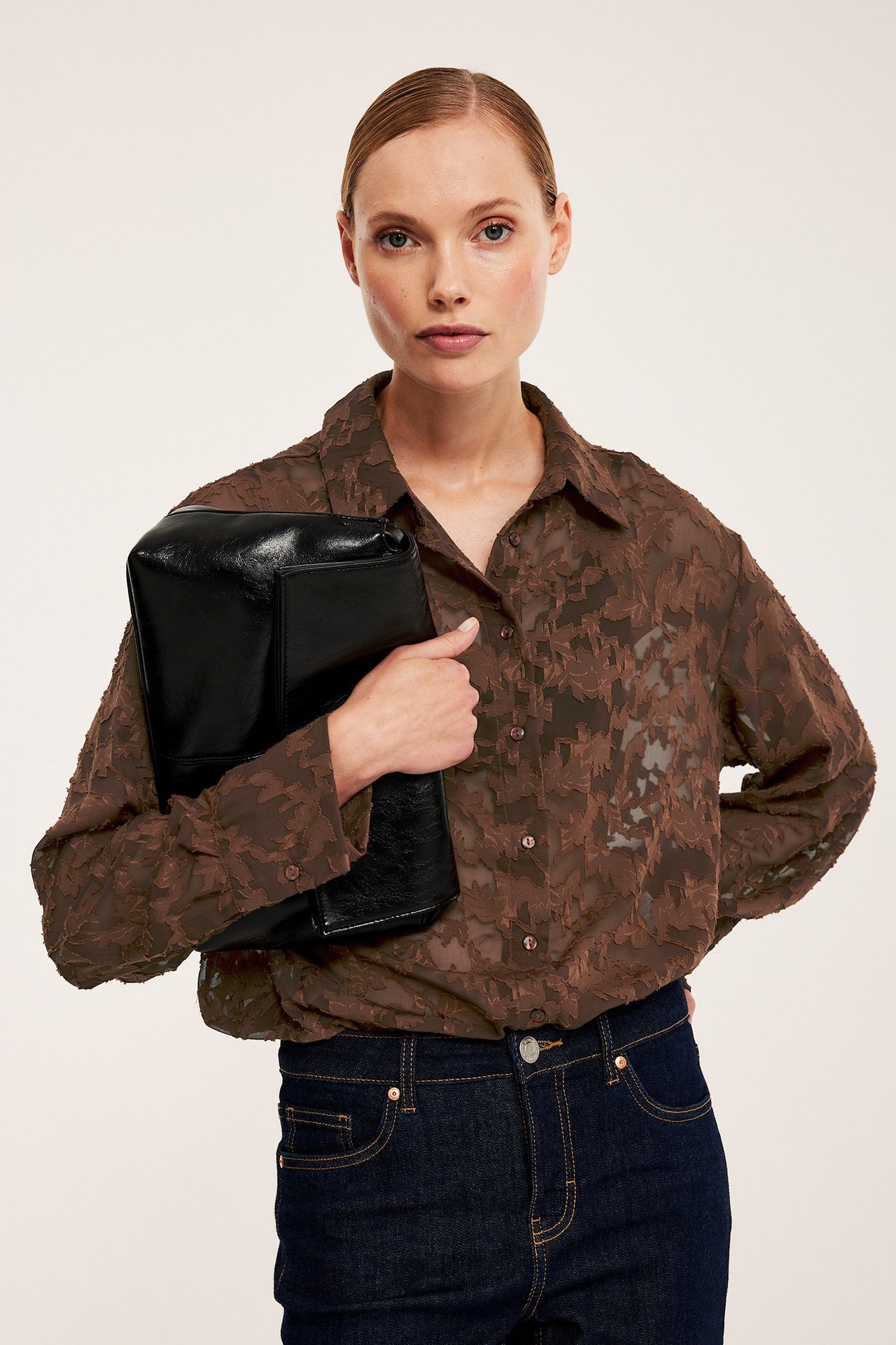 Framifrån: kvinna i brun transparent skjorta med jacquardmönster, mörka jeans och svart kuvertväska.