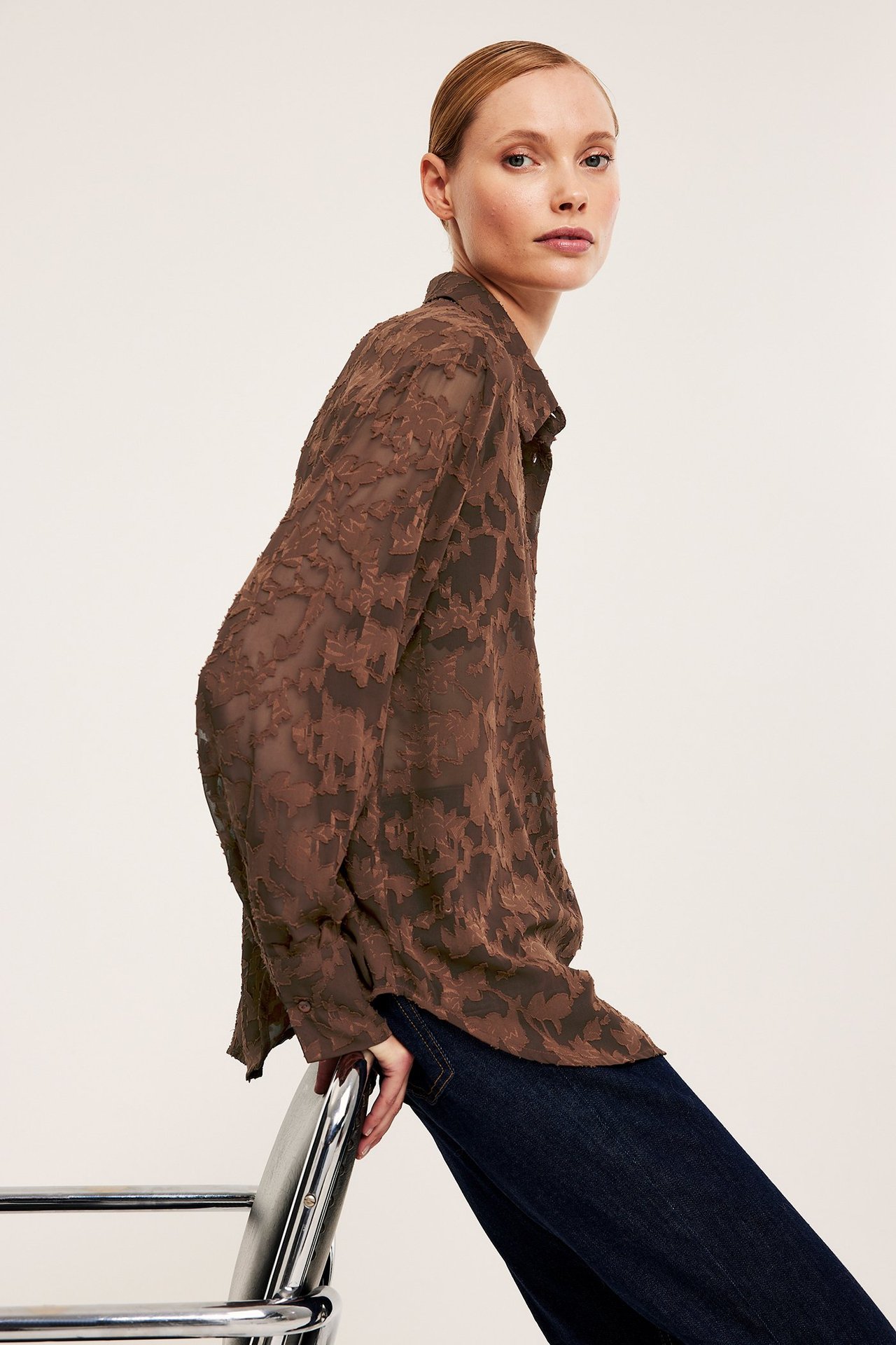 Kvinna i sidovy bär en brun jacquardmönstrad blommönstrad skjorta och mörkblå jeans.