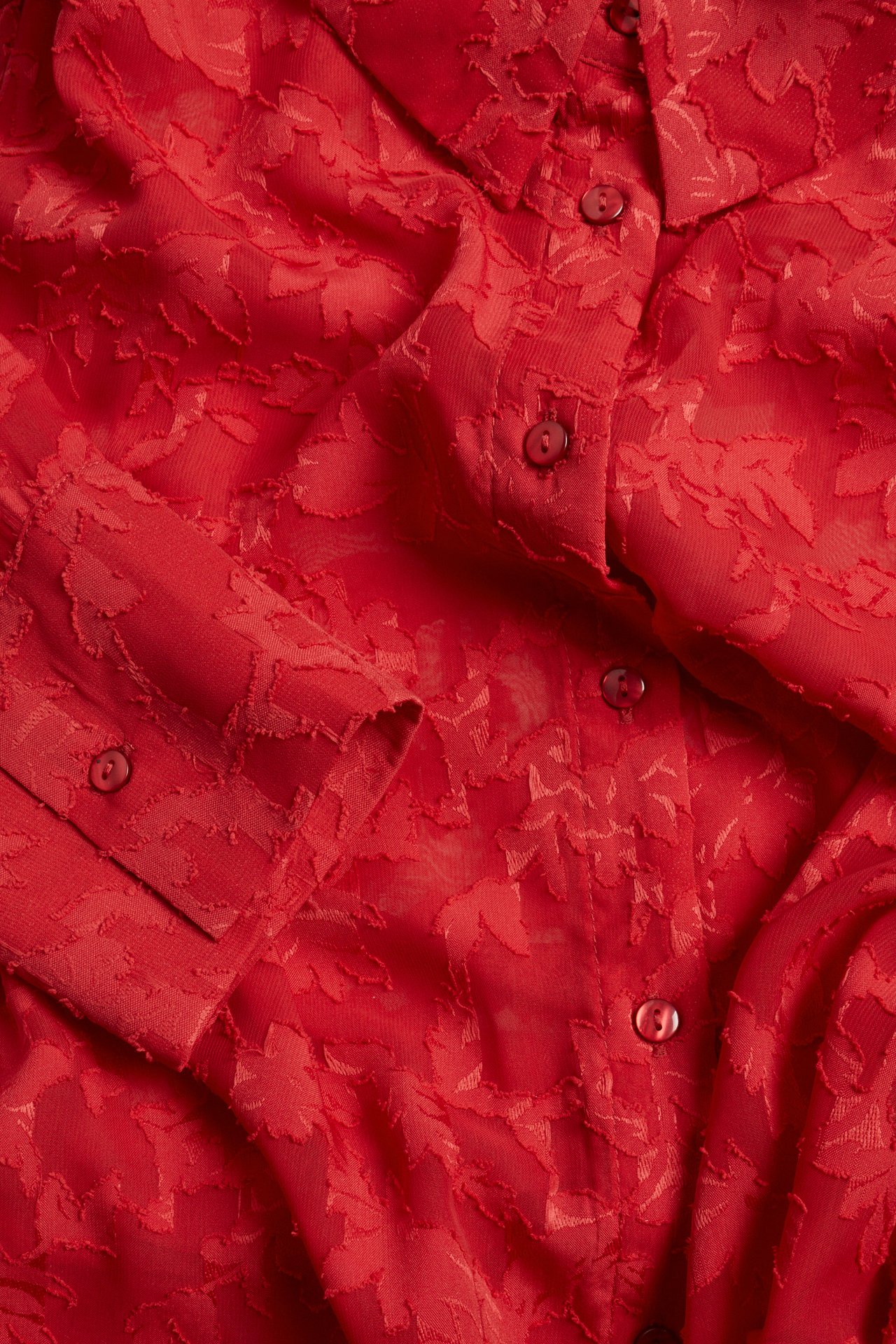 Zbliżenie, widok z góry, czerwonej koszuli z trójwymiarowym kwiatowym wzorem żakardowym, zapinanej na guziki i mankiety.