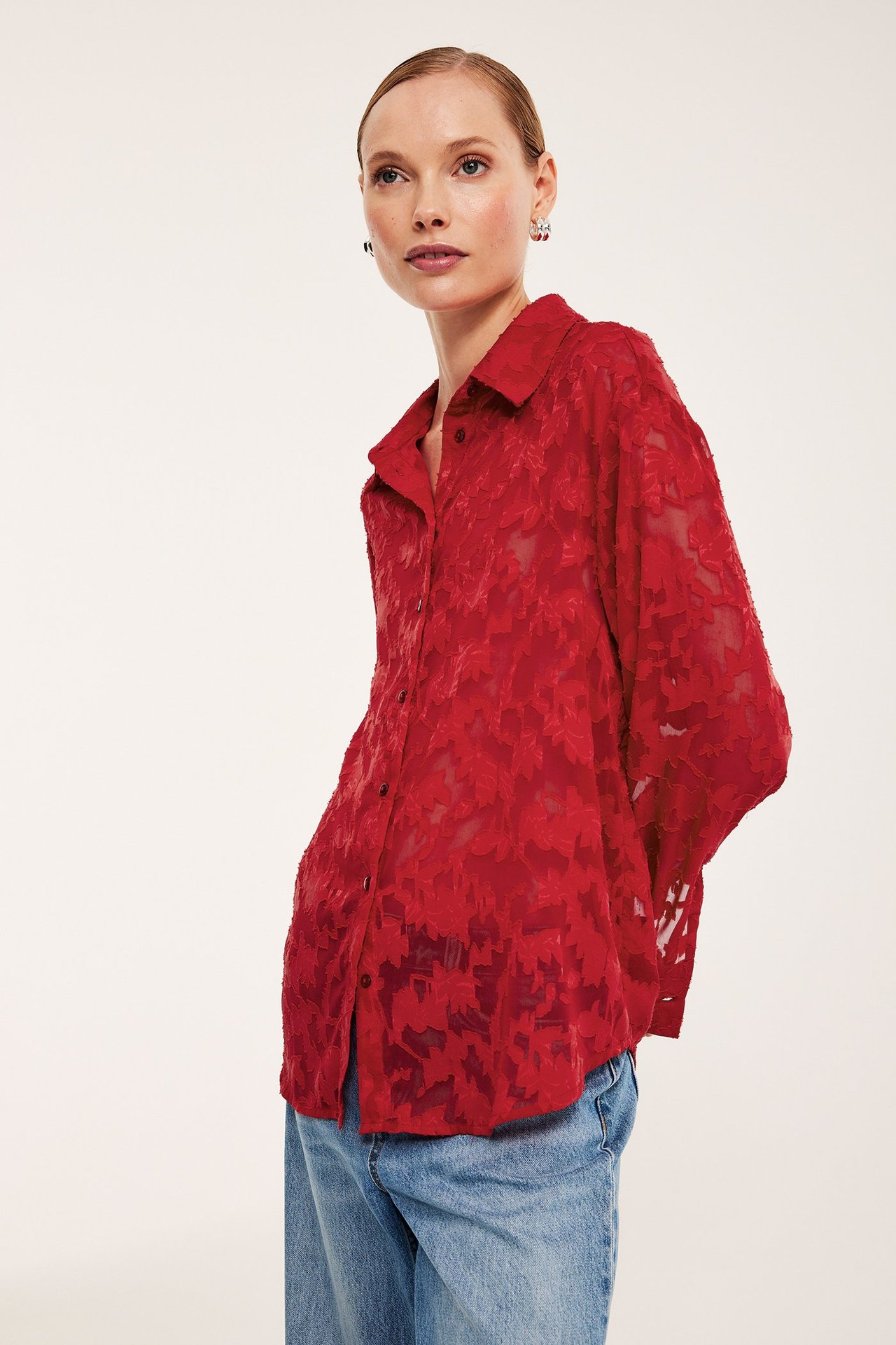 Z przodu: Kobieta w czerwonej luźnej koszuli żakardowej w kwiaty z guzikami, do tego niebieskie dżinsy.