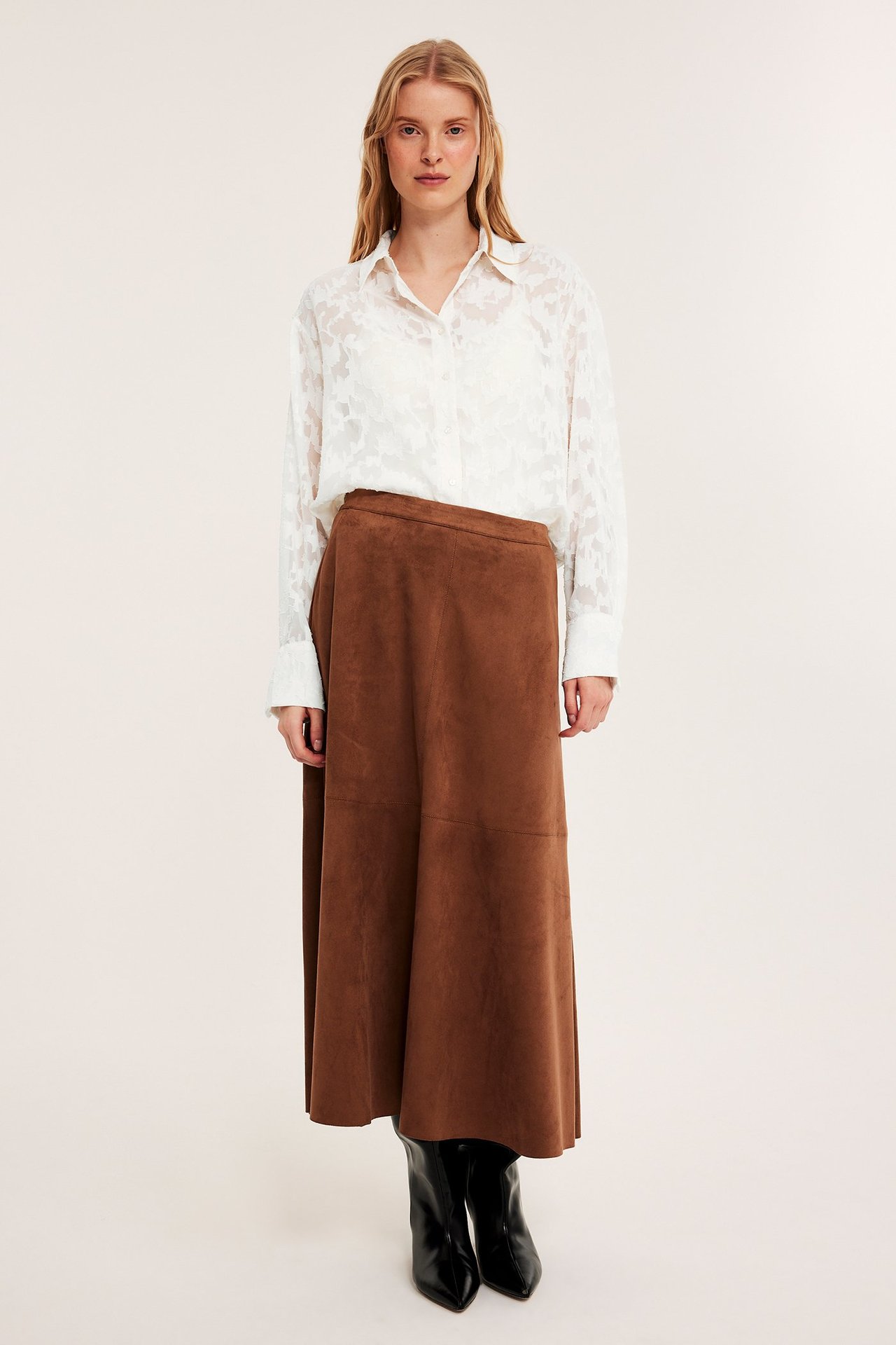 Nainen edestäpäin yllään luonnonvalkoinen jakardikuvioinen paita, ruskea liehuhelmainen hame ja mustat saappaat.