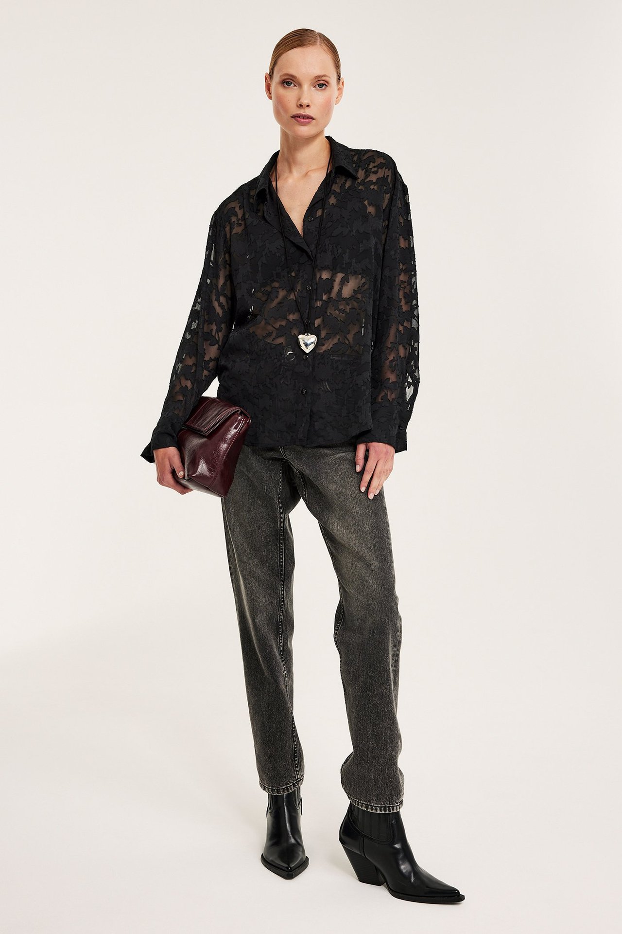Framifrån: Kvinna i svart blommig jacquardskjorta, grå jeans, svarta stövlar, hållande röd handväska.