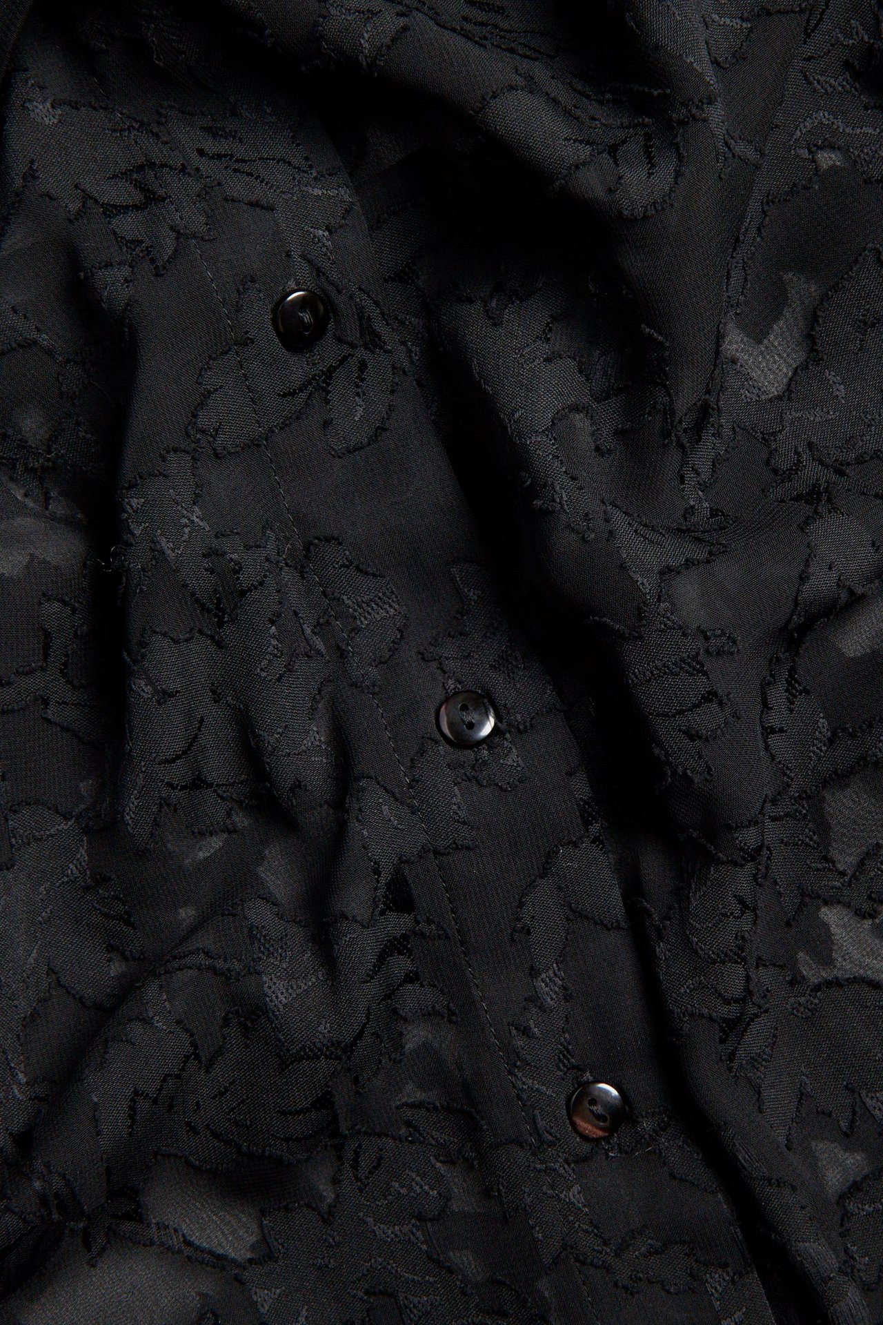 Zbliżenie czarnej koszuli z poliestru żakardowego z wytłoczonym wzorem kwiatowym i czarnymi guzikami.