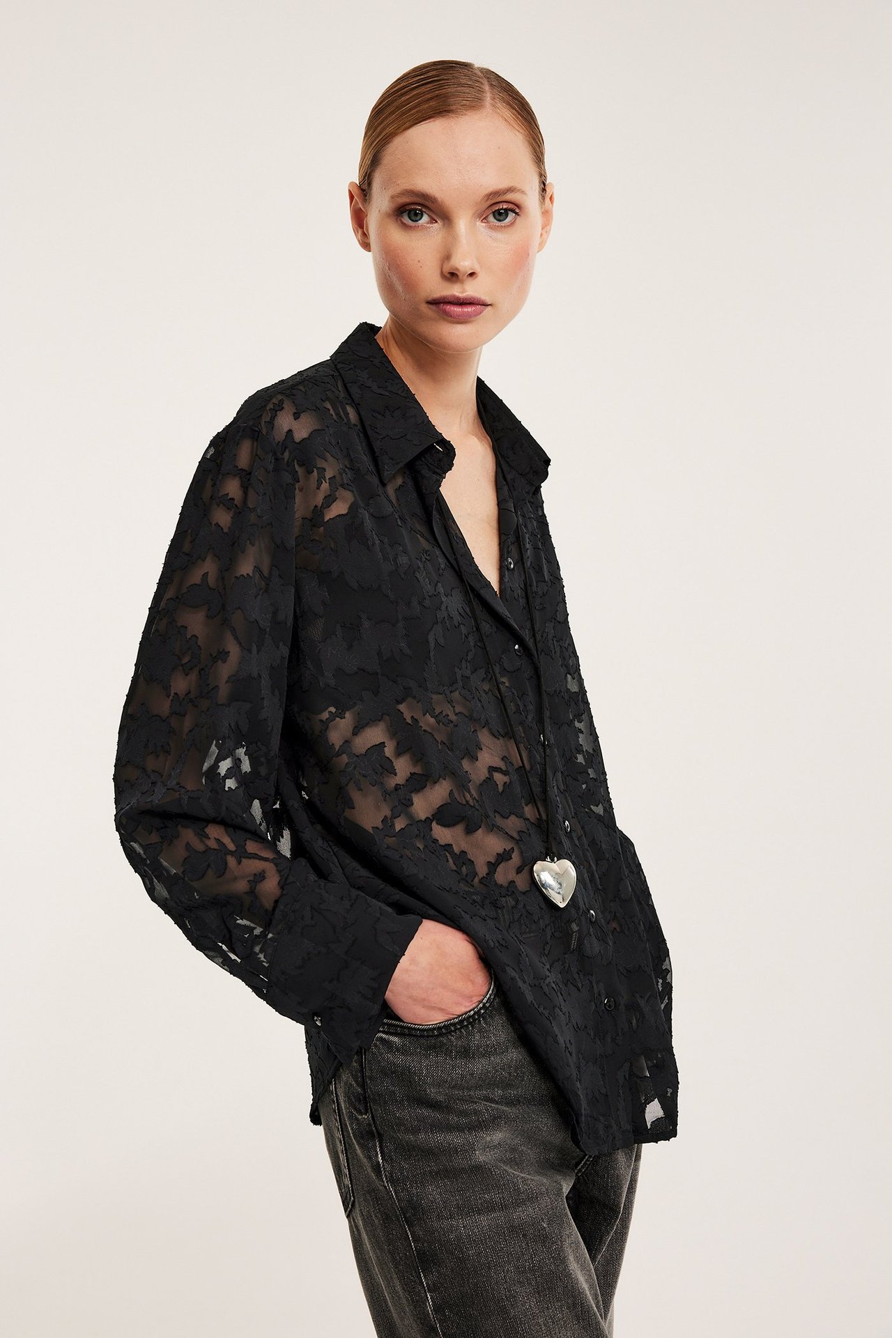Framifrån, kvinna i svart lös blommönstrad jacquard-skjorta, mörkgrå jeans och silverfärgat hjärthalsband.