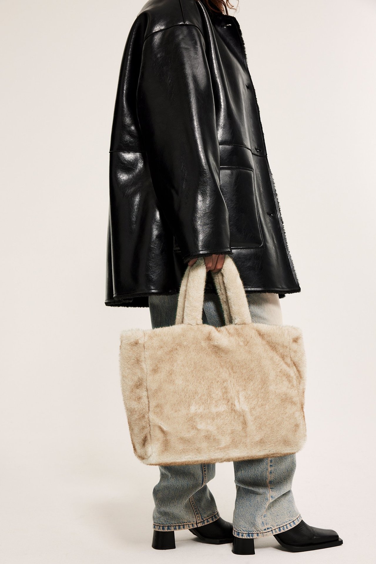 Widok z boku kobiety trzymającej beżową torbę ze sztucznego futra, w czarnej kurtce i niebieskich dżinsach.