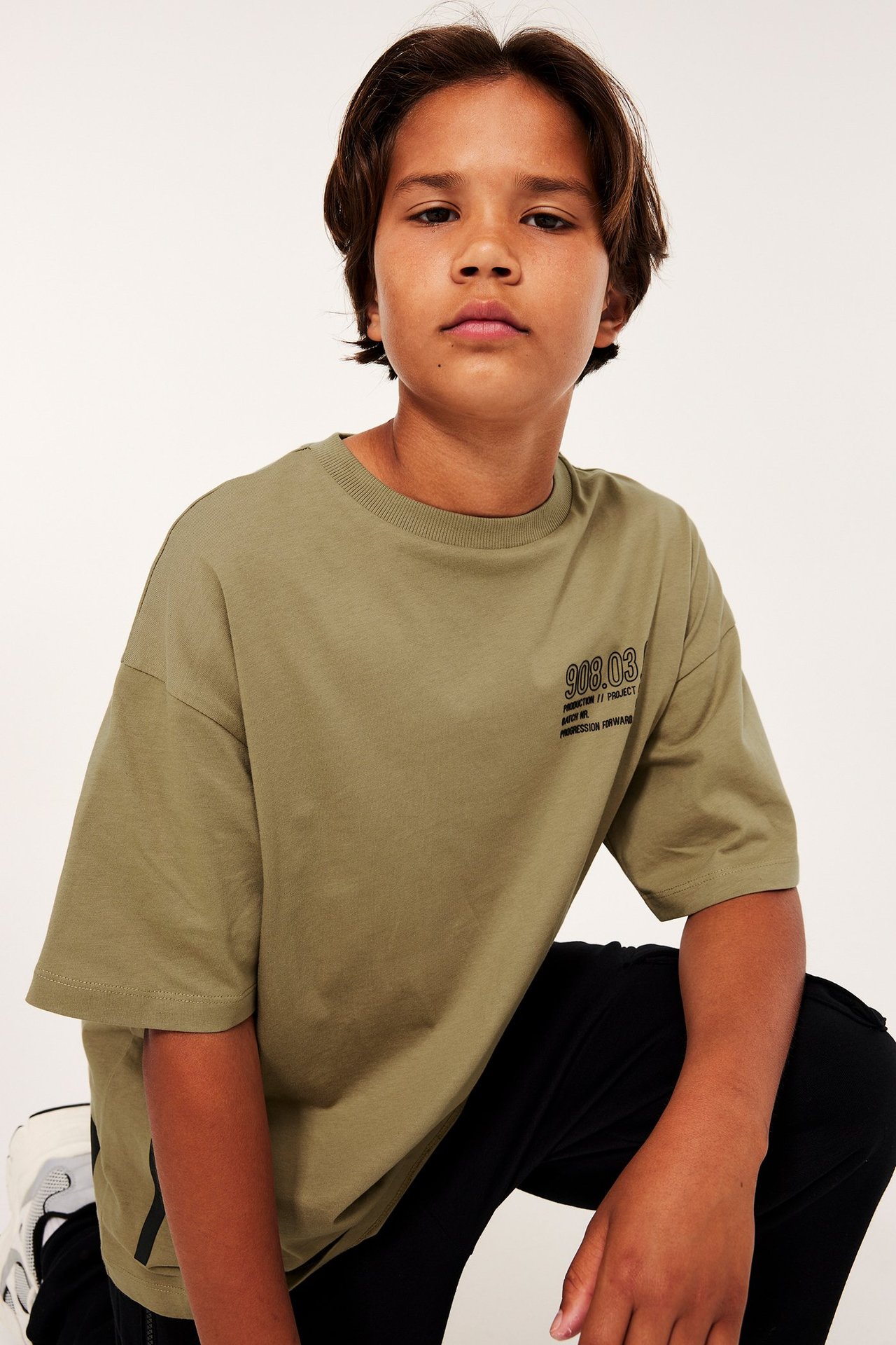 Widok z przodu dziecka w luźnym bawełnianym T-shircie khaki z nadrukiem, czarnych spodniach i sneakersach.