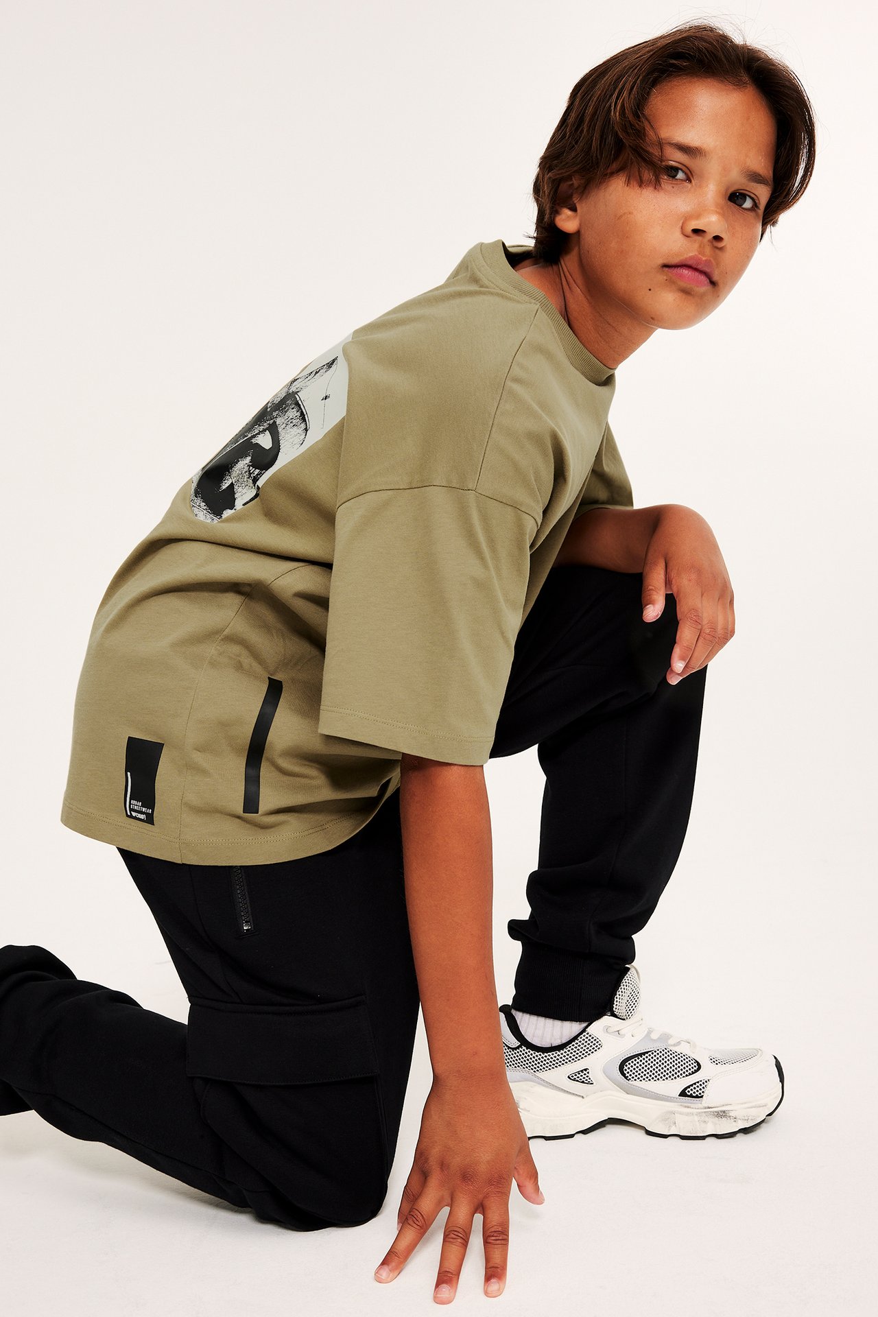Sidovy av ett barn i en olivgrön oversized T-shirt med ryggtryck, svarta cargobyxor och vita sneakers.