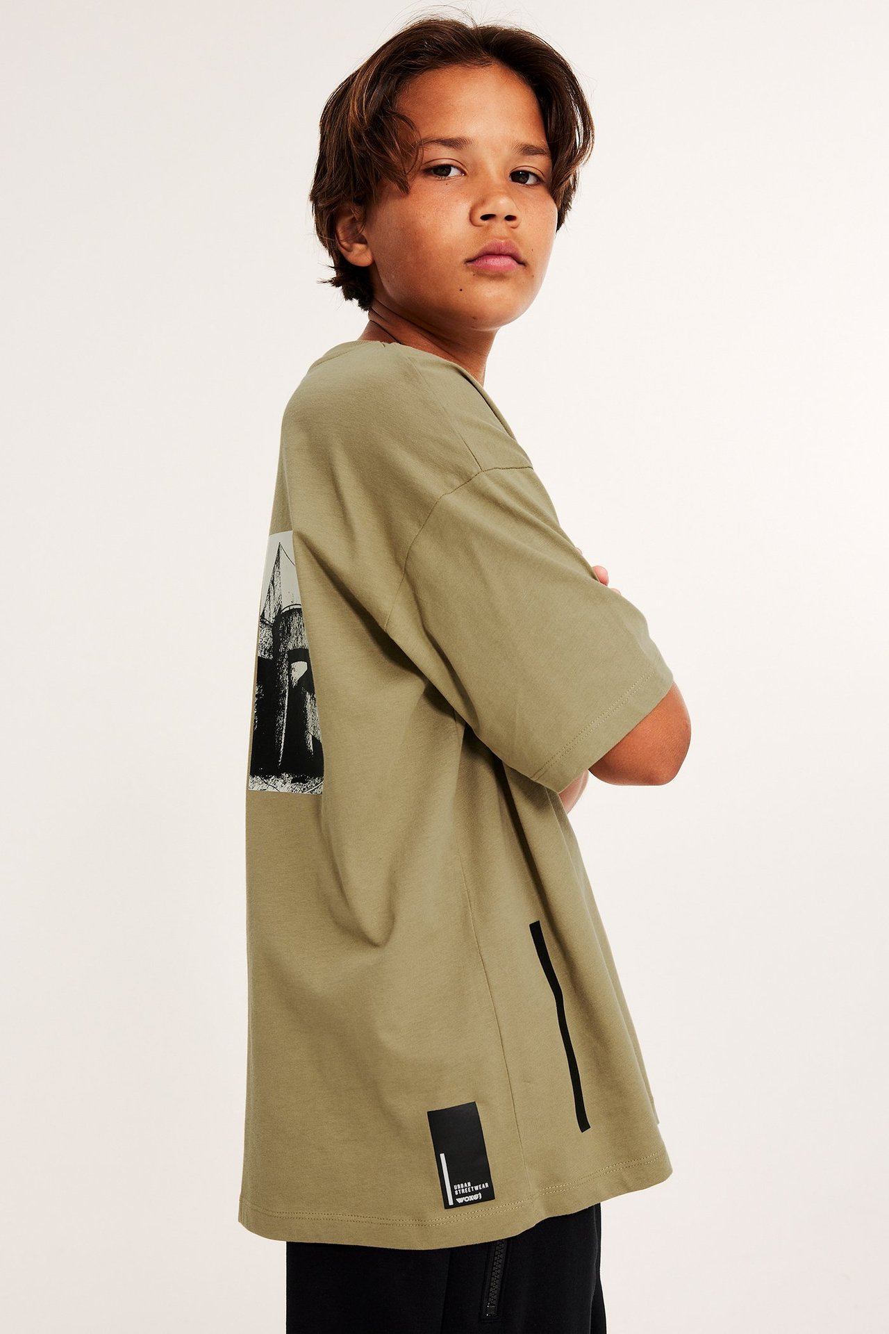 Widok z boku dziecka w luźnym bawełnianym T-shircie khaki z dużym nadrukiem i czarnych spodniach.