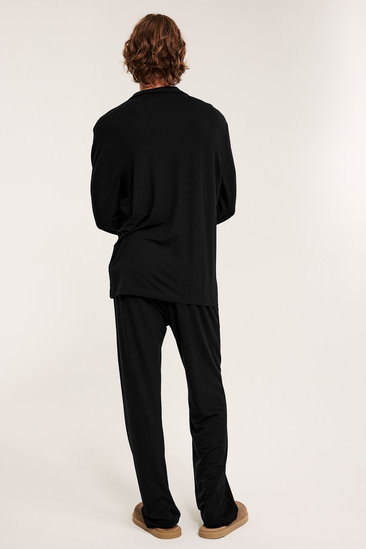 Mies takaapäin mustassa, löysässä pyjamasettiin, jossa paita ja housut, yllään ruskeat tohvelit.