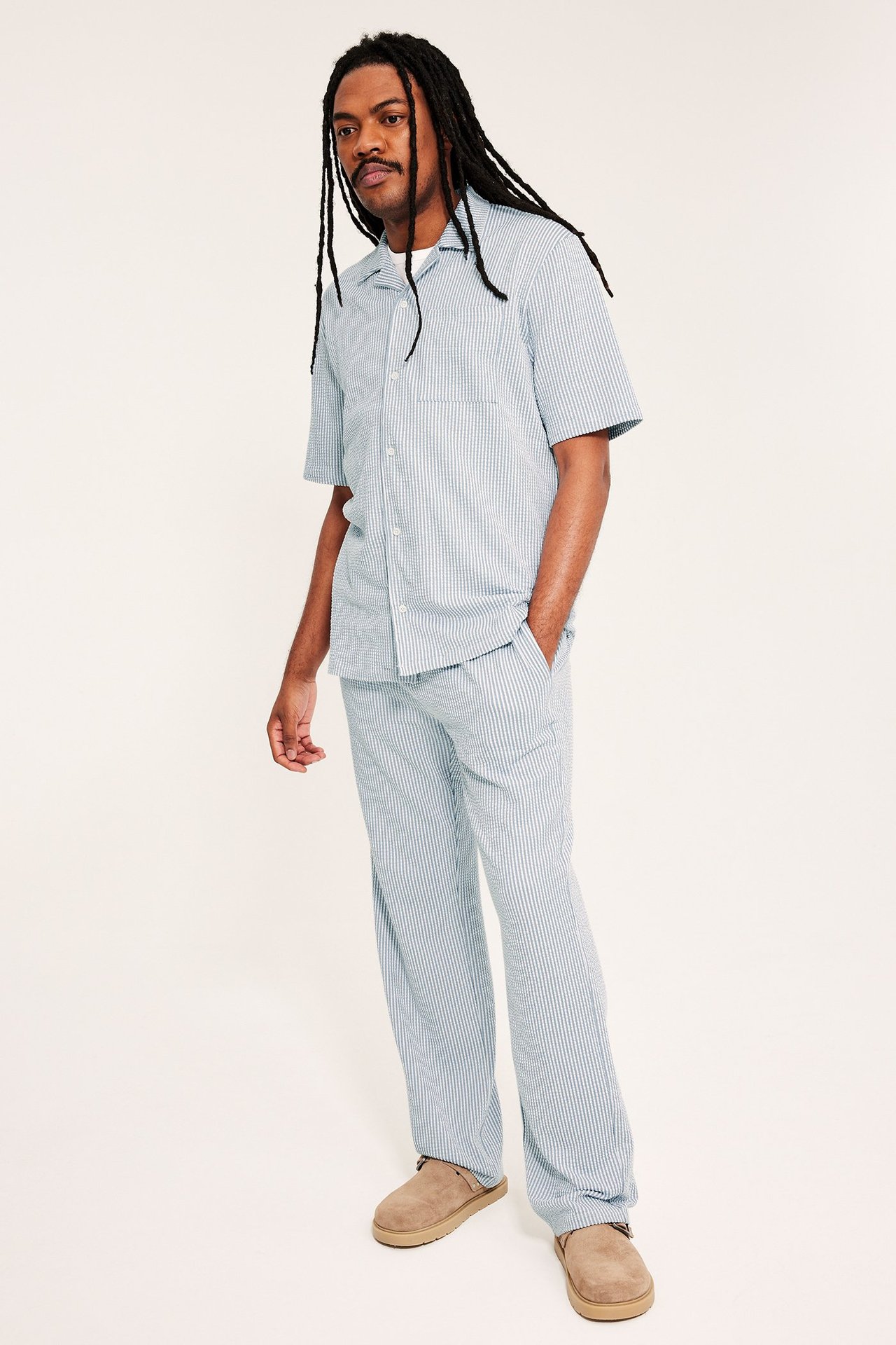 Edestä: mies vaaleansinisissä raidallisissa seersucker-pyjamahousuissa, yhteensopivassa lyhythihaisessa paidassa ja ruskeissa tohveleissa.