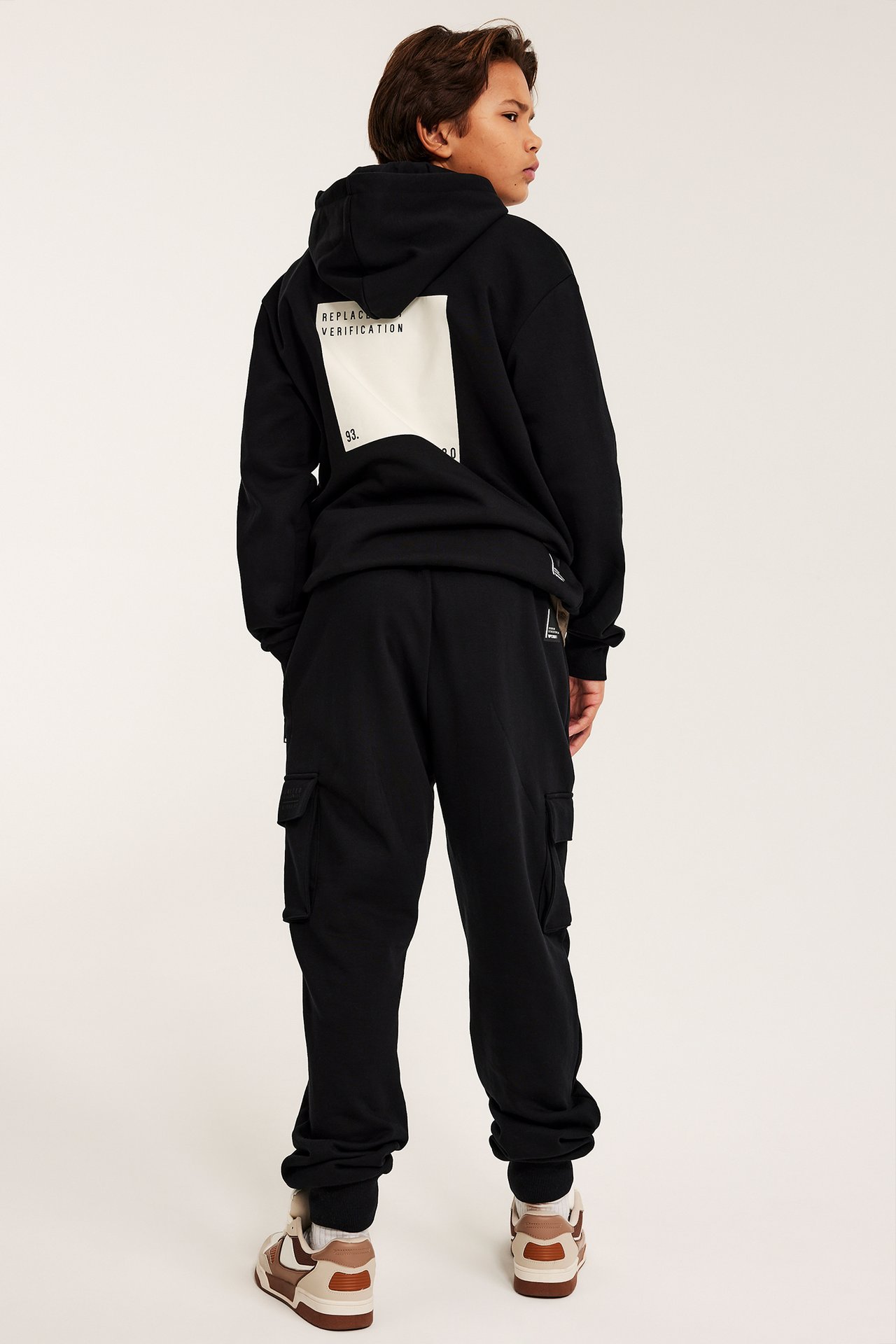 Widok z tyłu dziecka w czarnych bojówkach dresowych i czarnej bluzie z jasnym nadrukiem na plecach.