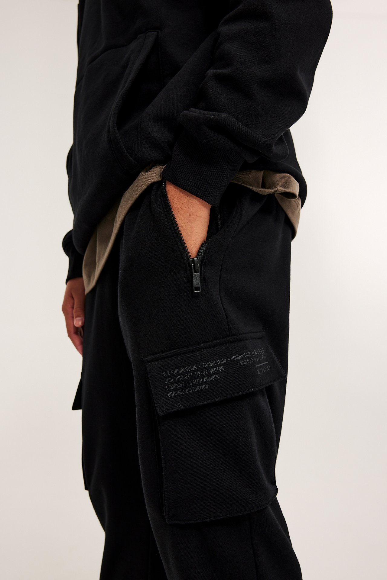 Widok z boku: czarne dresy bawełniane z zamkiem i kieszenią cargo z nadrukiem, noszone przez osobę.