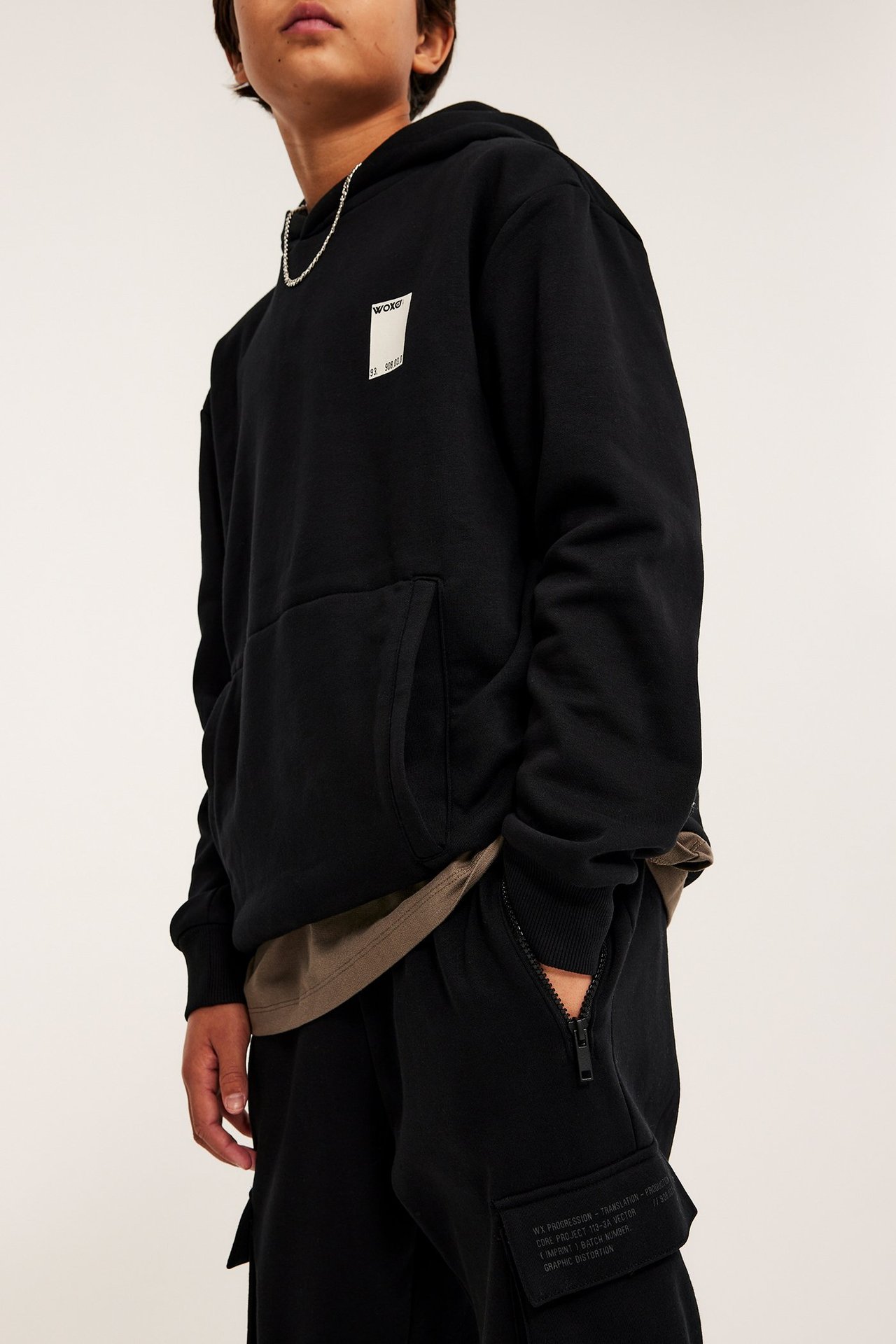 Framifrån, kid i svart tryckt huvtröja, svarta mjukisbyxor med dragkedjeficka och silverkedja.