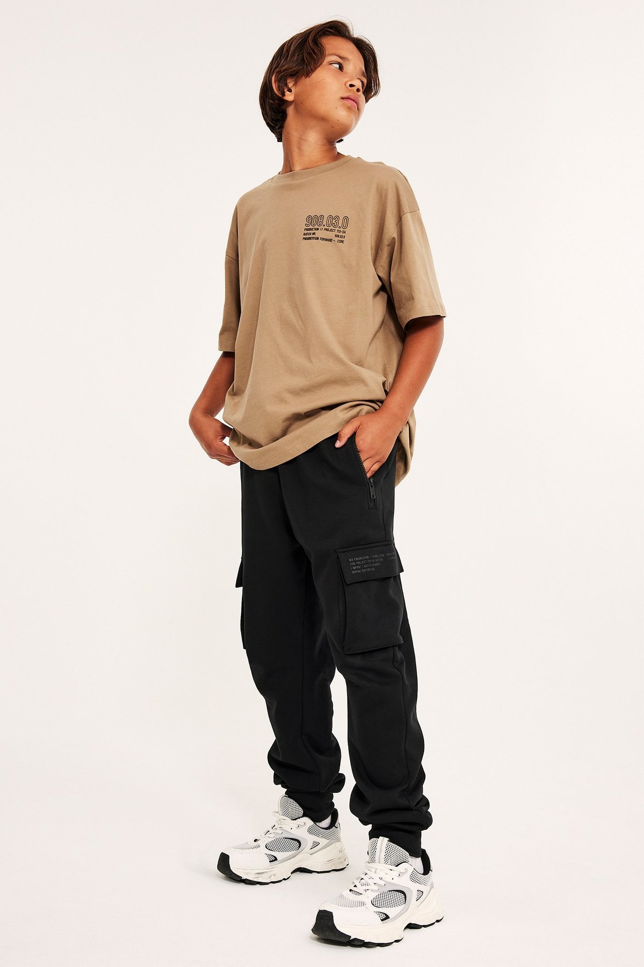 Widok z przodu dziecka w czarnych spodniach dresowych cargo, beżowej koszulce z nadrukiem i białych sneakersach.