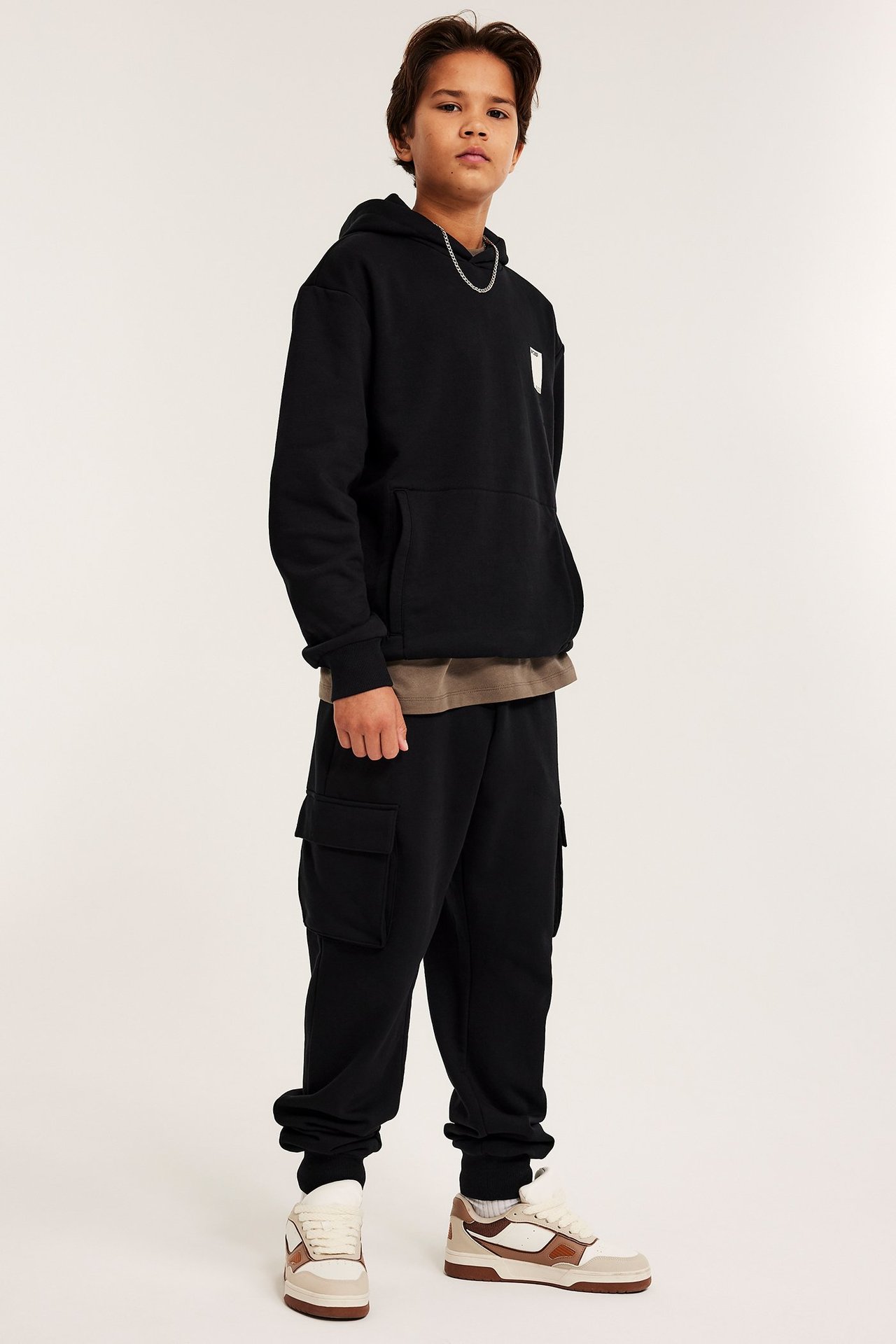 Framifrån, ett barn i svarta cargomjukisbyxor, svart hoodie, brun t-shirt, silverkedja och sneakers.