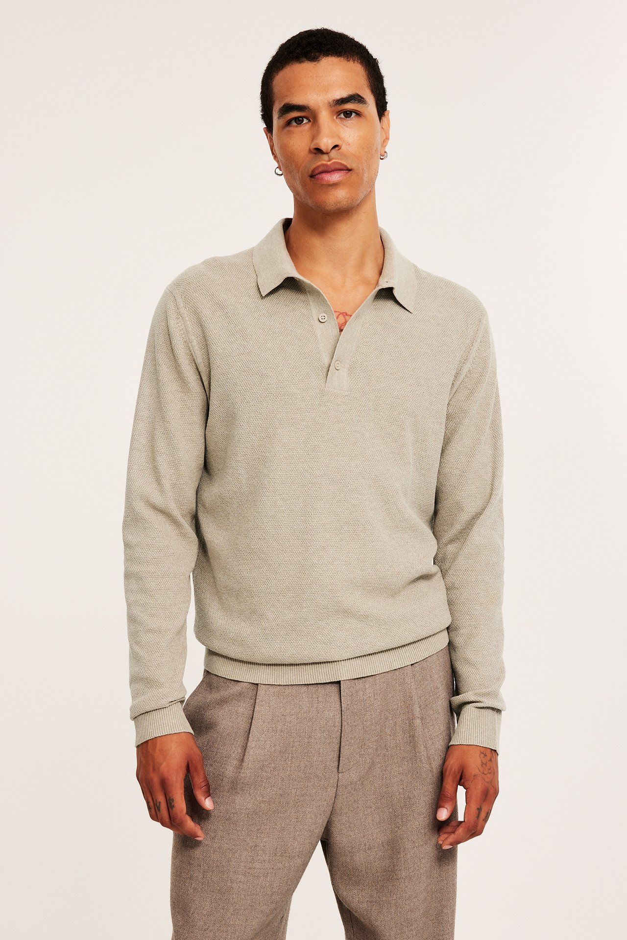 Frontbilde av en mann i beige melert strukturstrikket pikétrøye med to knapper og brune plisserte bukser.