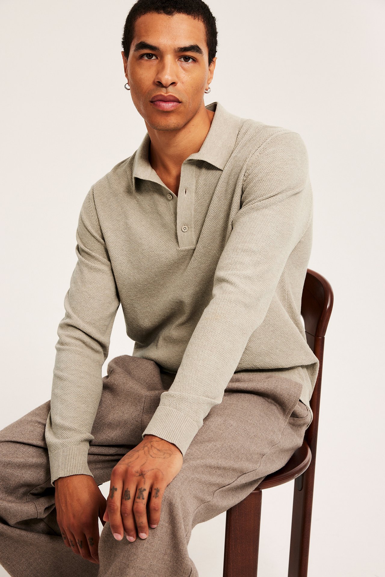 Frontbilde av en mann i en beige melert strukturstrikket pologenser med knapper, sammen med brune melert bukser.