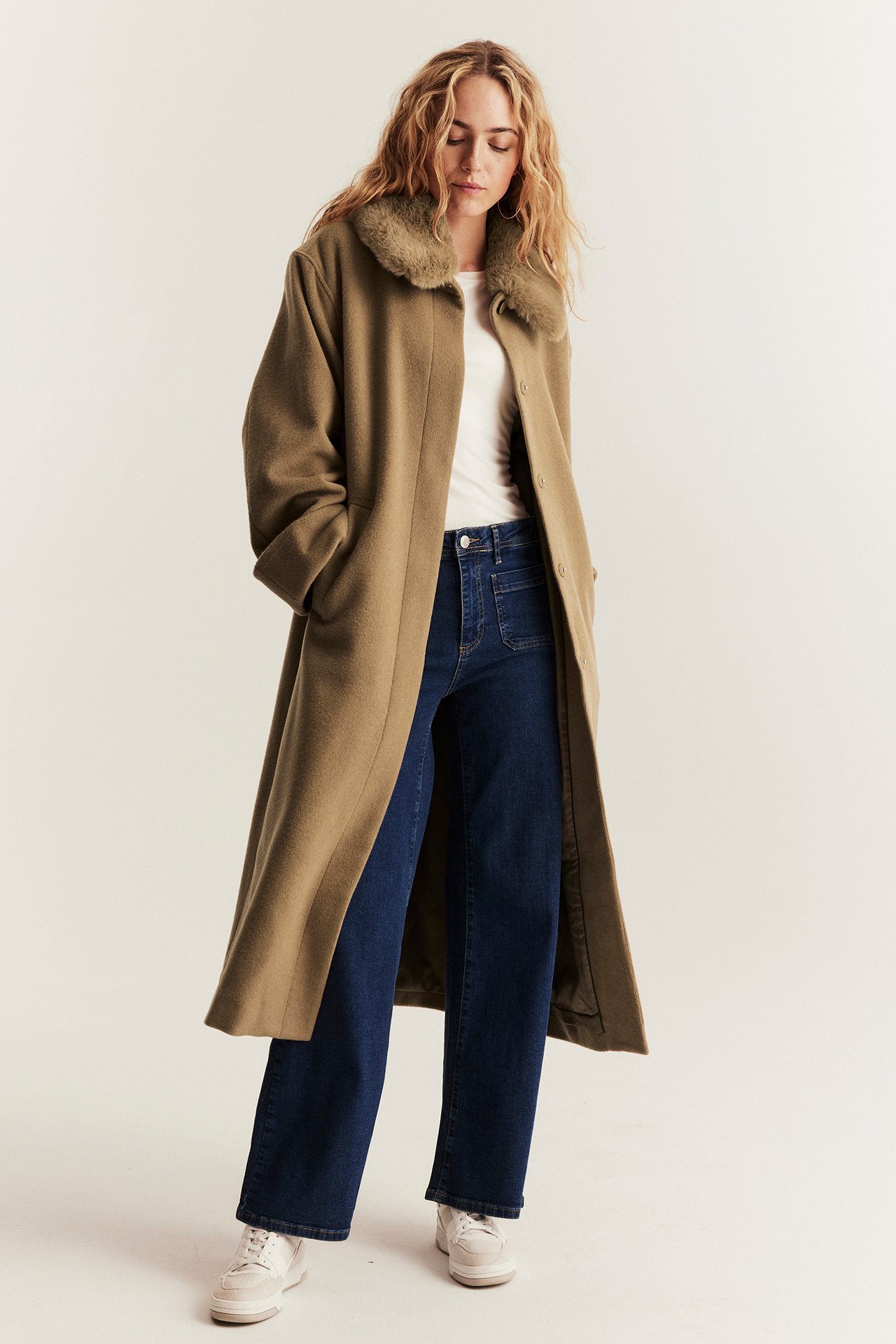 Widok z przodu: Kobieta nosi otwarty brązowy płaszcz wełniany z futrzanym kołnierzem, biały t-shirt i ciemnoniebieskie dżinsy.