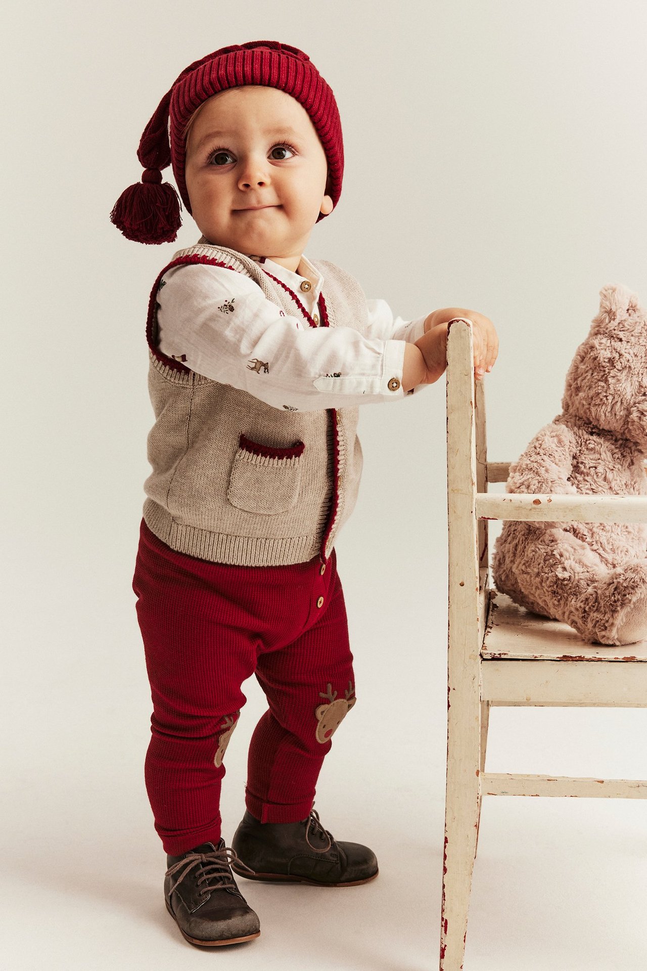 Barn i røde leggins med reinsdyrapplikasjon, beige vest, hvit skjorte, rød lue.