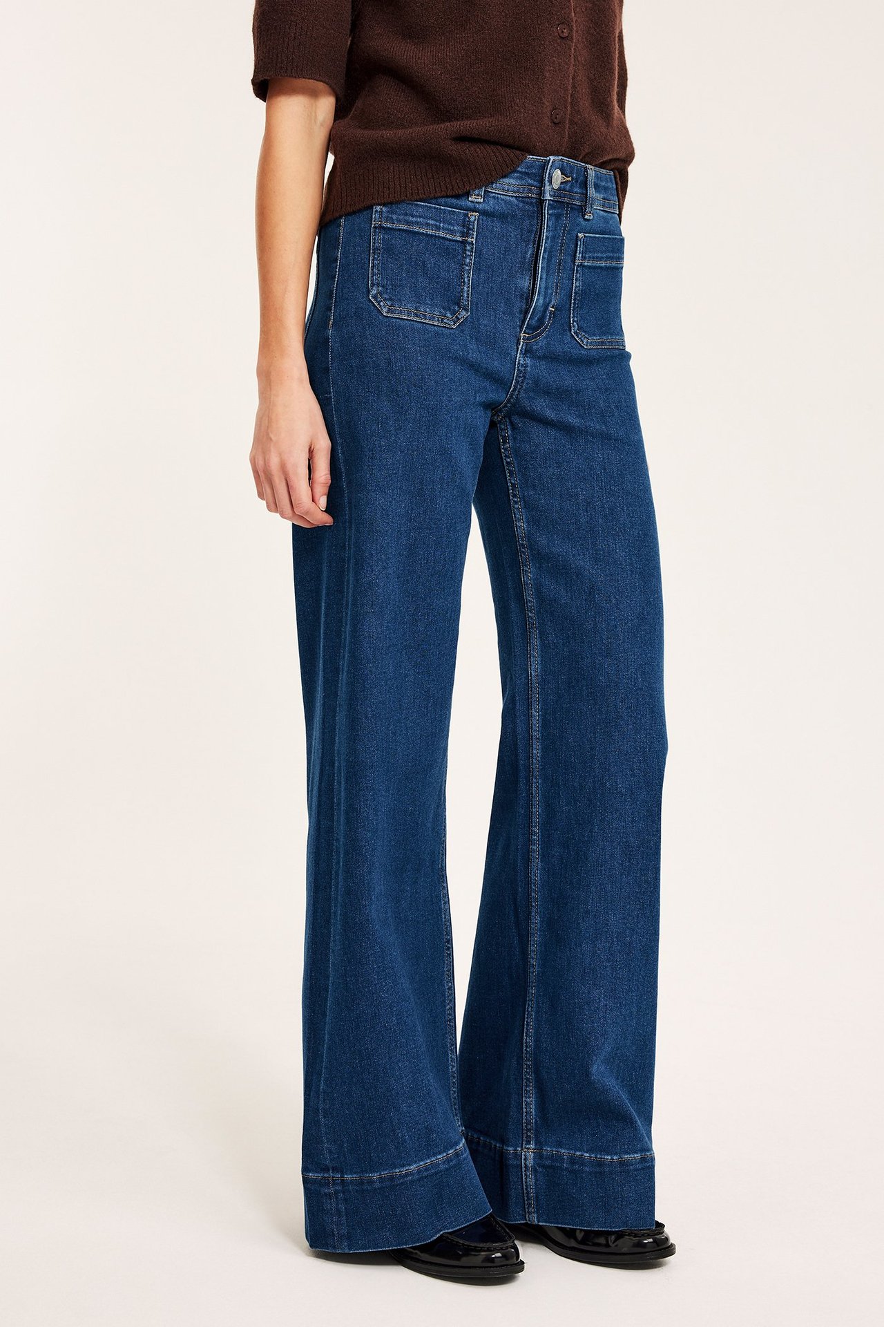 Frontvisning av kvinne i blå denim vide jeans med frontlommer, stylet med brun topp og svarte loafers.