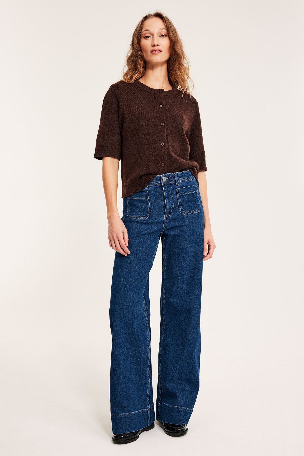 Widok z przodu: Kobieta w szerokich dżinsach high-waist z kieszeniami, brązowym krótkim kardiganie i czarnych butach.