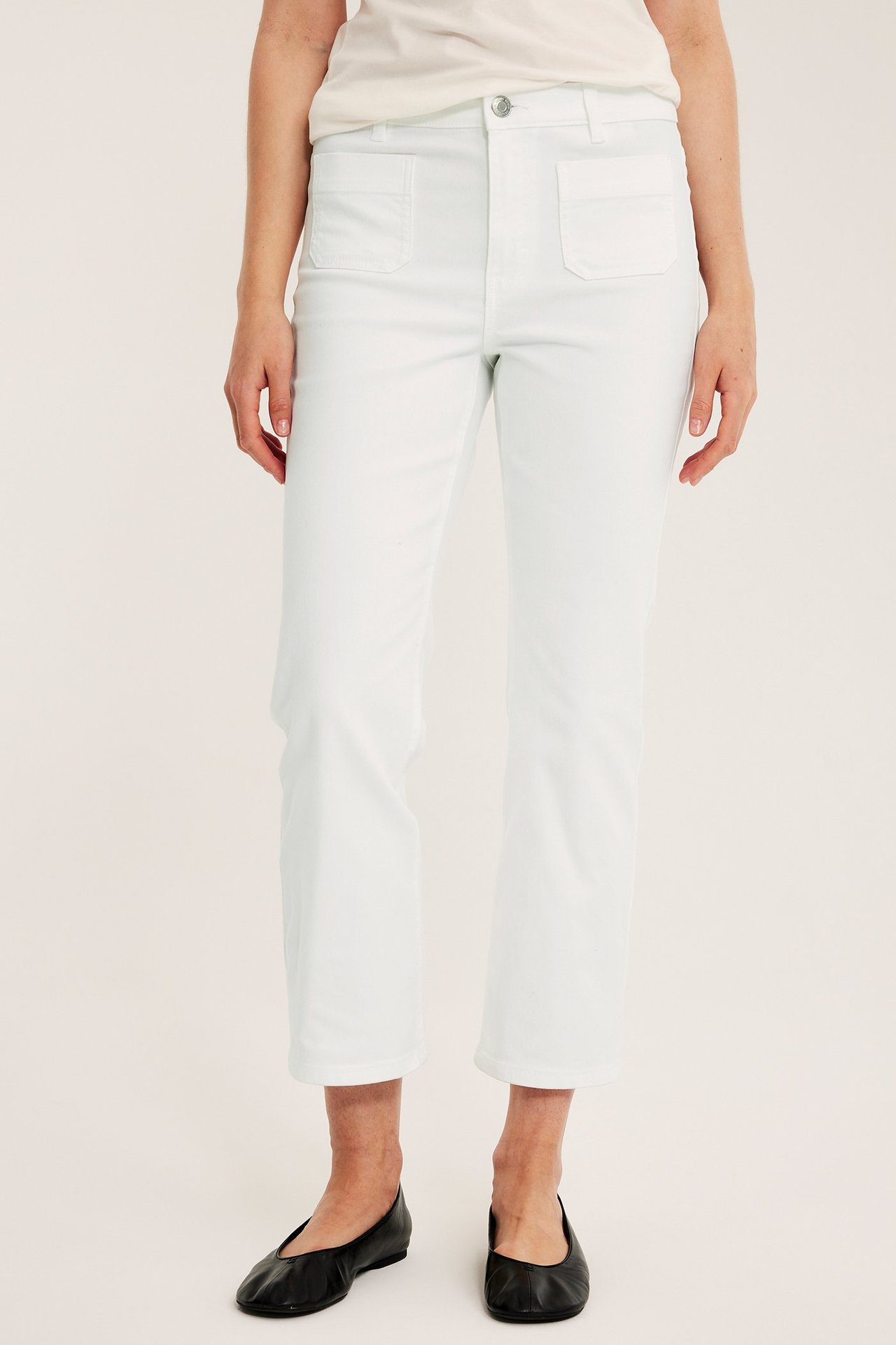Framifrån: Kvinna i vita croppade flare-jeans med frontfickor, en vit t-shirt och svarta ballerinaskor.