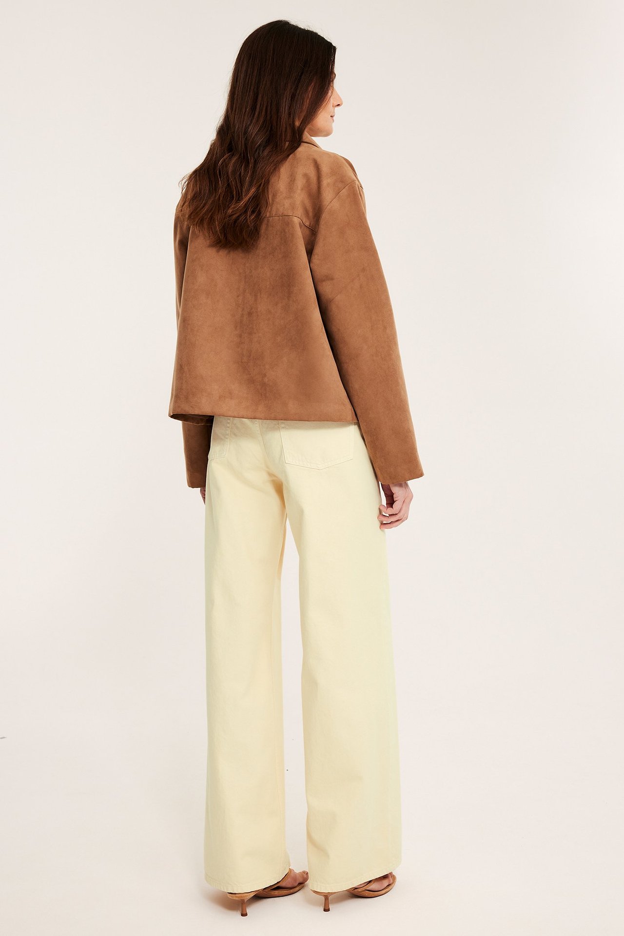 Sned bakifrån-vy: kvinna i ljusgula vida twilljeans, brun jacka och bruna klackskor.