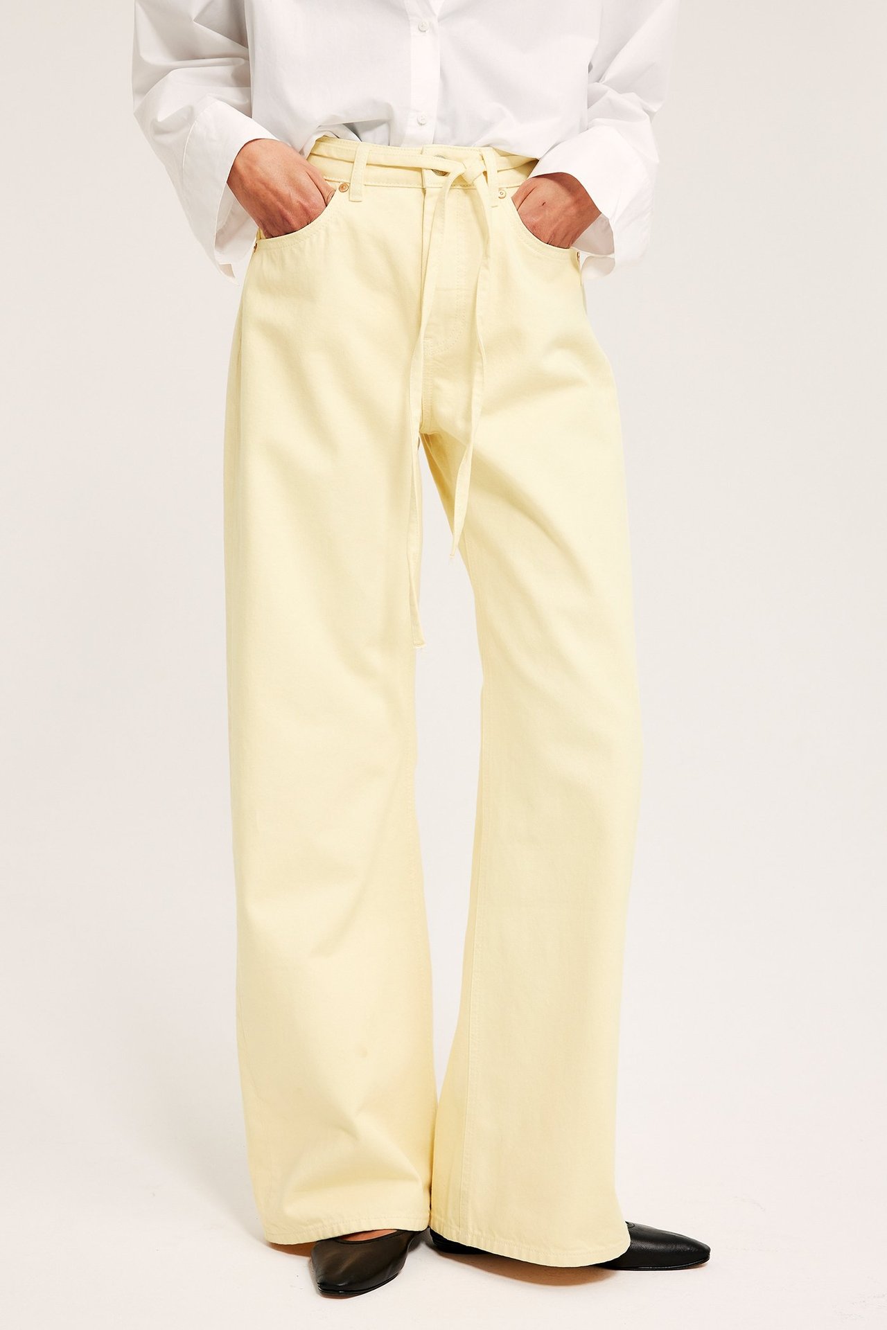 Framifrån: Kvinna i ljusgula vida 5-ficksjeans med knytskärp, vit skjorta och svarta platta skor.