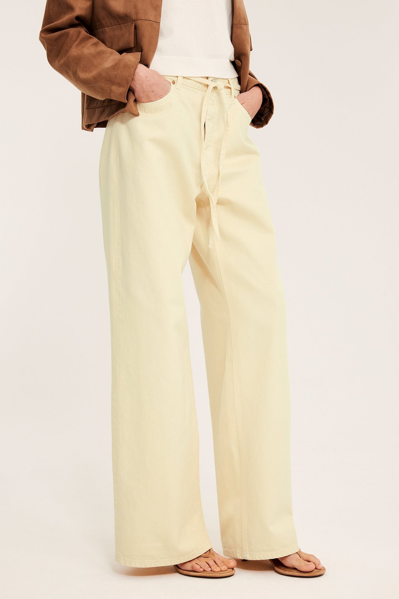 Forfra: kvinne i lysegule vide jeans med høy midje og snøring, hvit topp, brun jakke, sandaler.