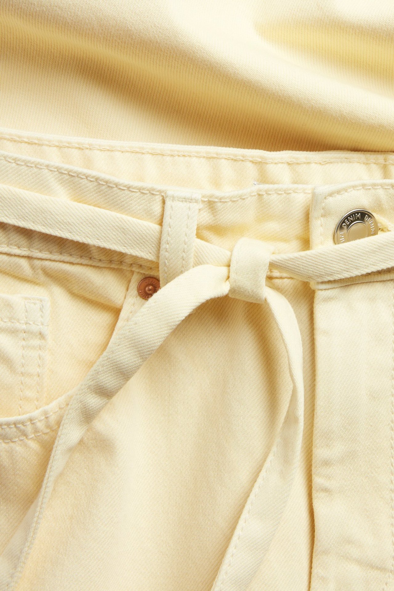Zbliżenie jasnożółtych dżinsów z szerokimi nogawkami: wiązany parciany pasek, pięć kieszeni, metalowe guziki.