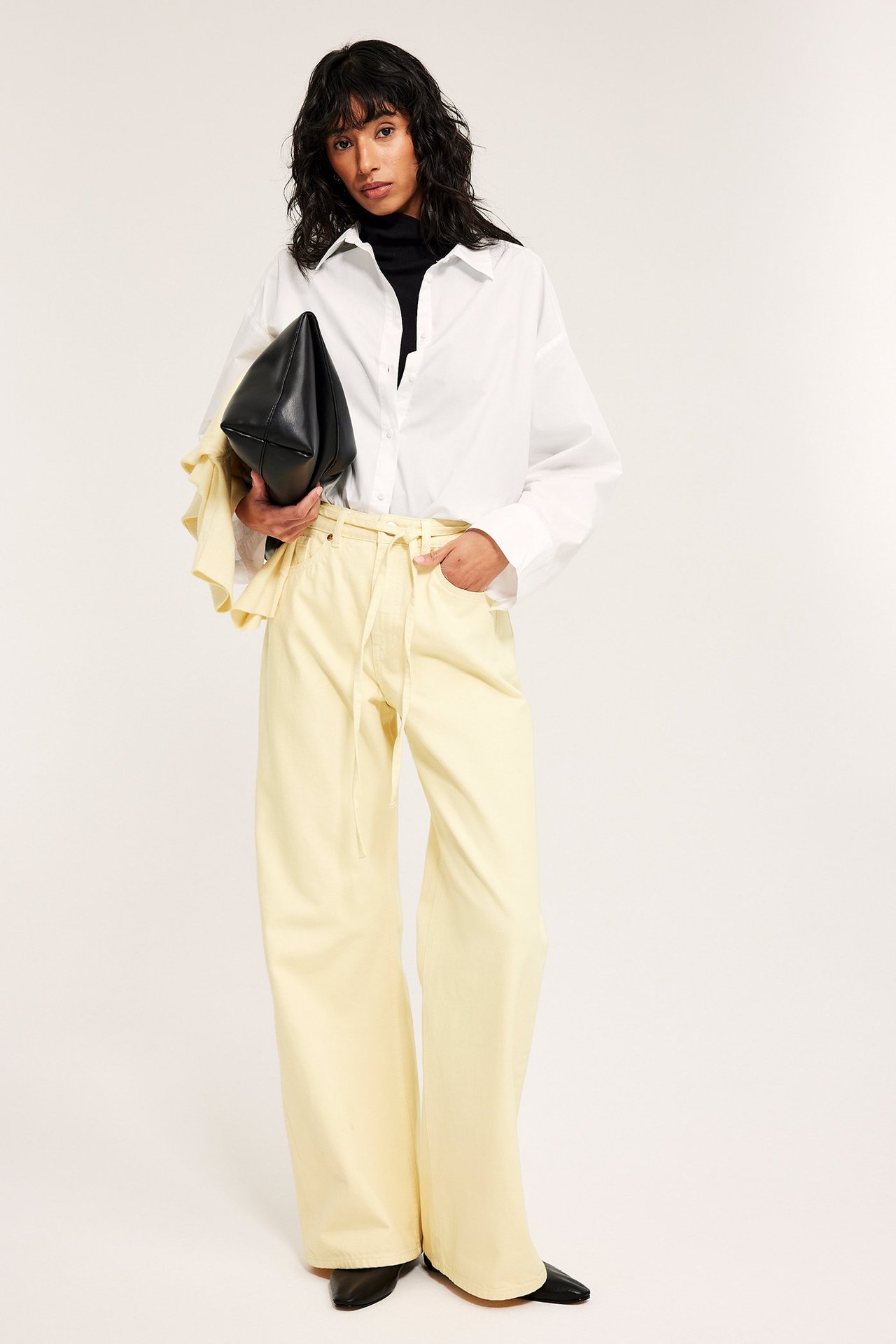 Frontvy: Kvinna i ljusgula vida jeans, vit skjorta, svart polotröja, håller en svart kuvertväska.