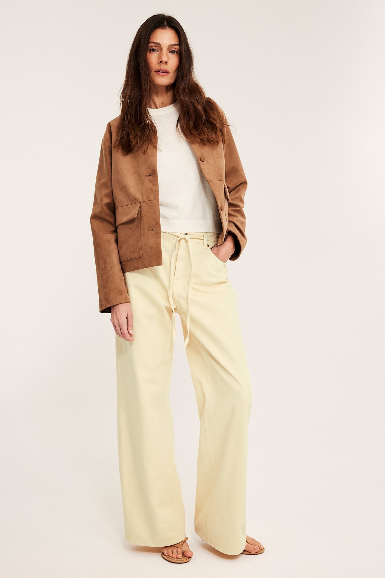 Framifrån, kvinna i ljusgula wide-leg jeans med dragsko, en vit topp och en brun kort jacka.