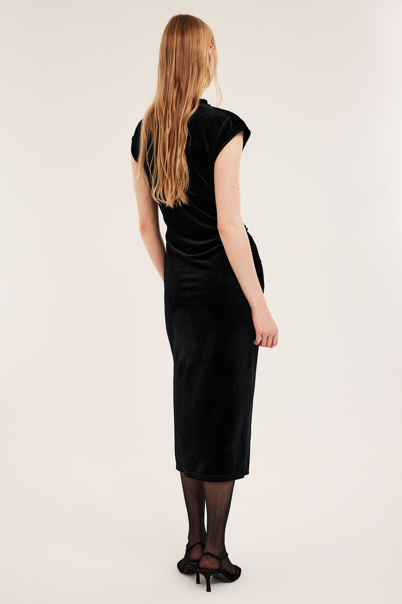 Ryggside av kvinne i en svart tettsittende fløyelskjole, tynne strømpebukser og svarte høyhælte sandaler.