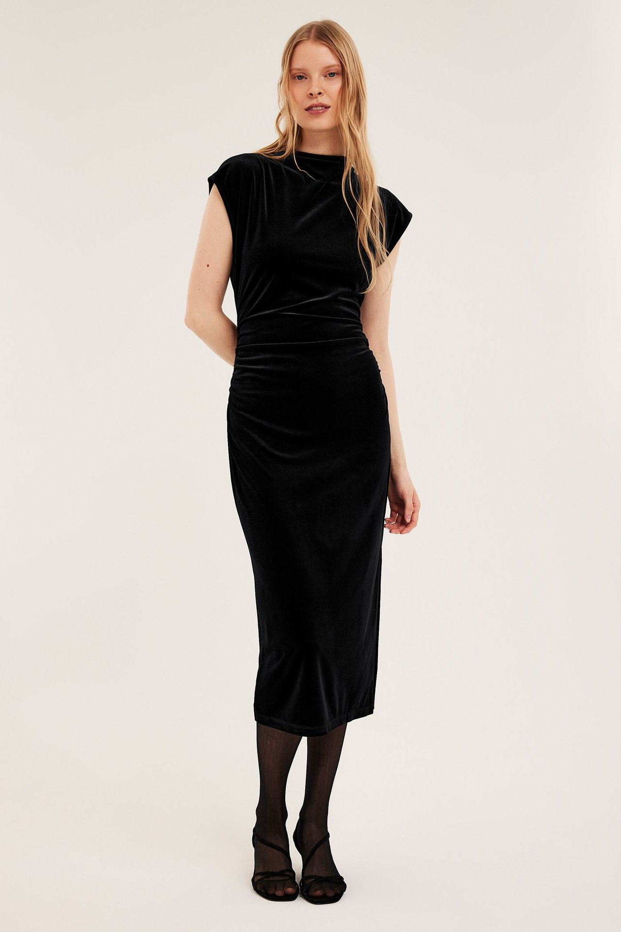 Forfra: Kvinne i tettsittende svart fløyelskjole med kortermer, rynker, sorte strømpebukser og sandaler.