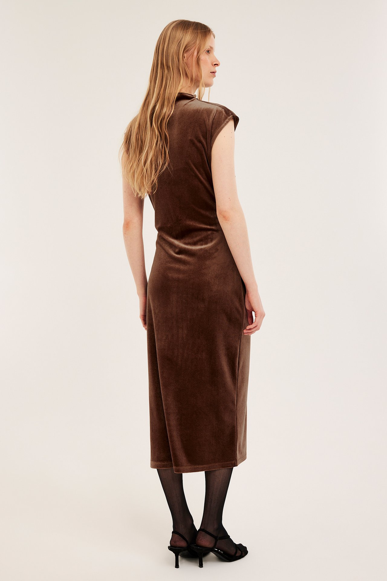 Boczny widok kobiety w obcisłej, brązowej aksamitnej sukience z rękawkiem, czarnych rajstopach i czarnych sandałach na obcasie.