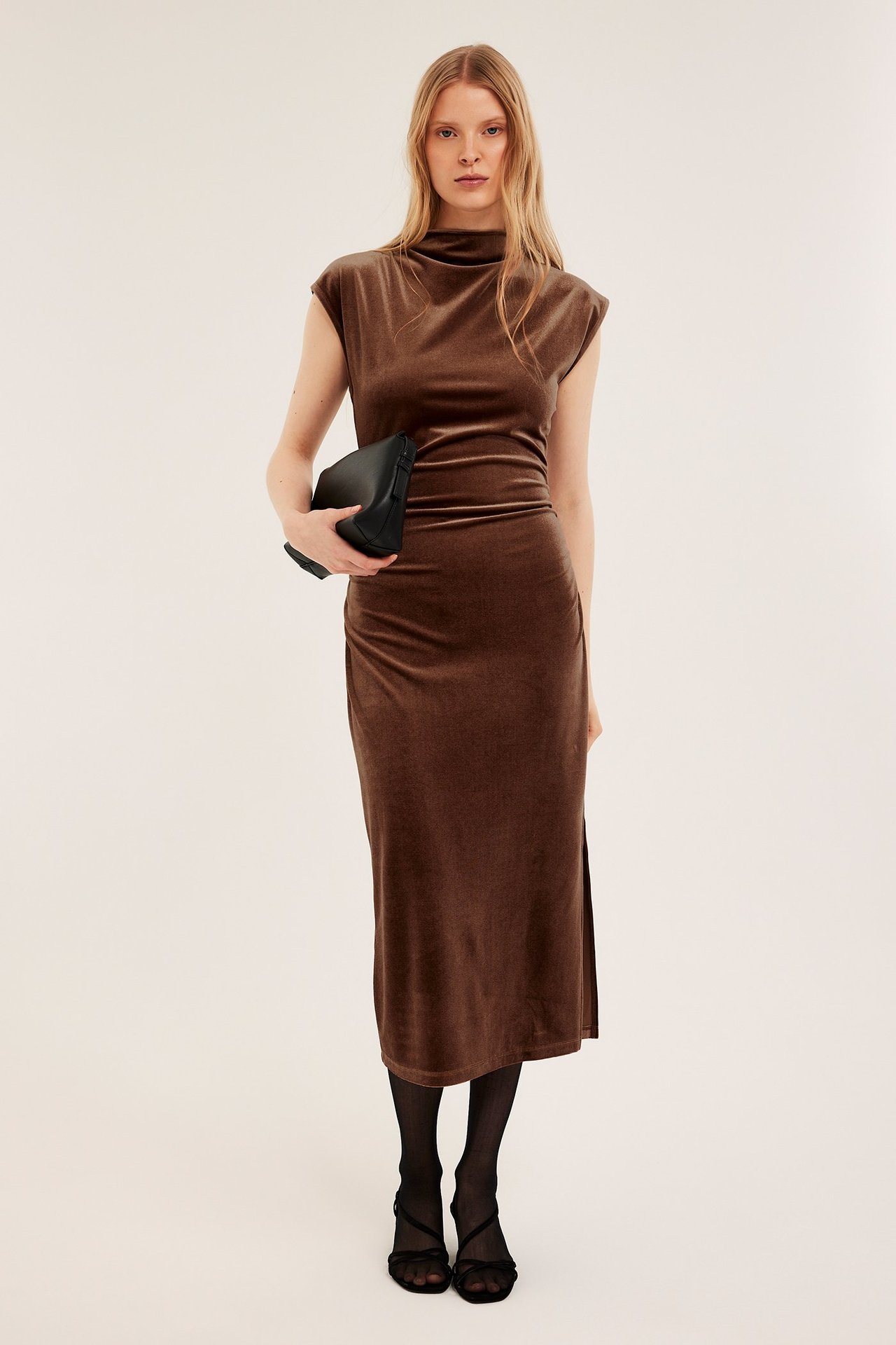 Front: kvinne i tettsittende brun fløyelskjole, korte ermer, holder svart clutch, svarte strømpebukser og hæler.