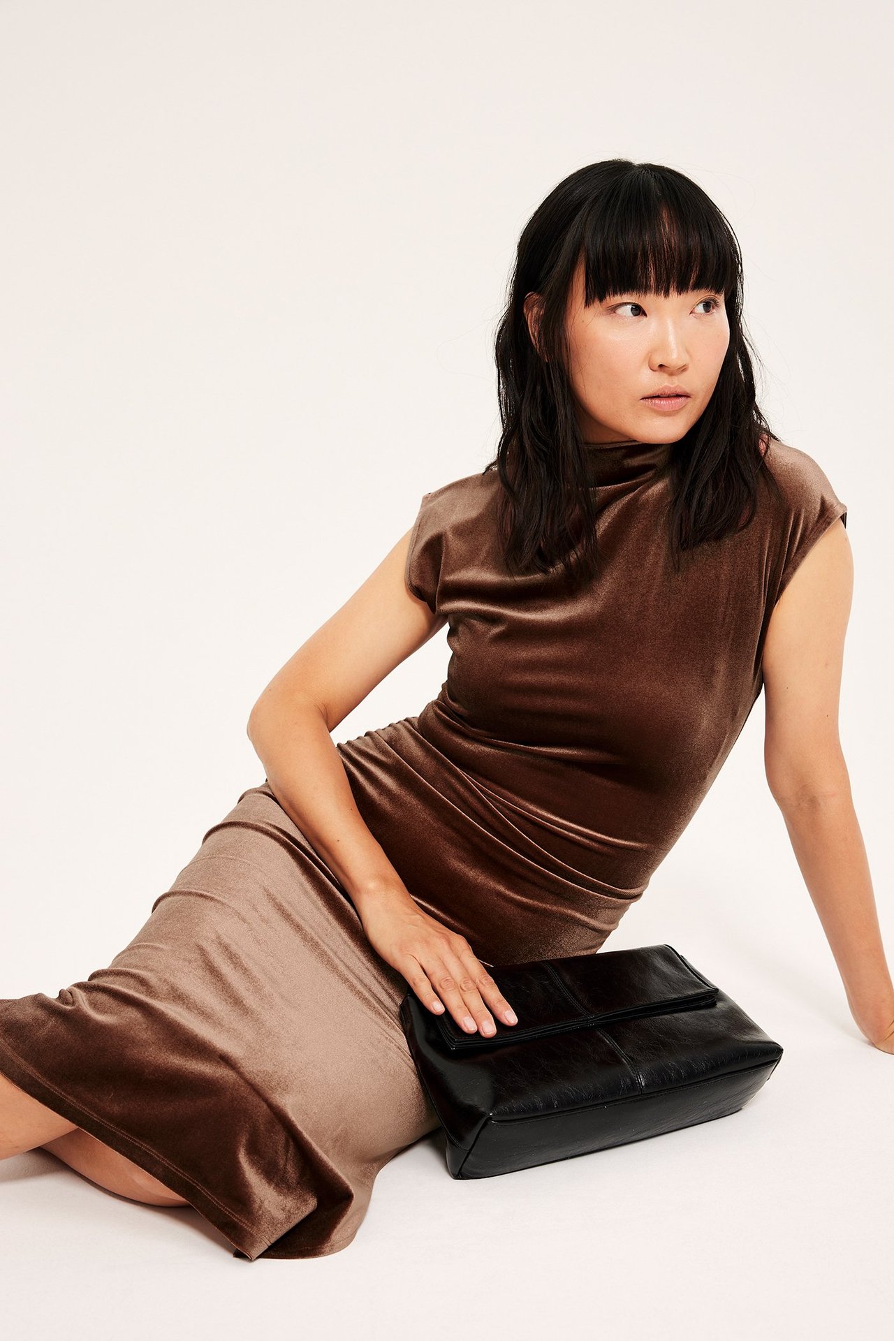 Nainen ruskeassa samettifarkkuvaatteessa istuu, edestä näkyvissä, ja pitää mustaa laukkua.