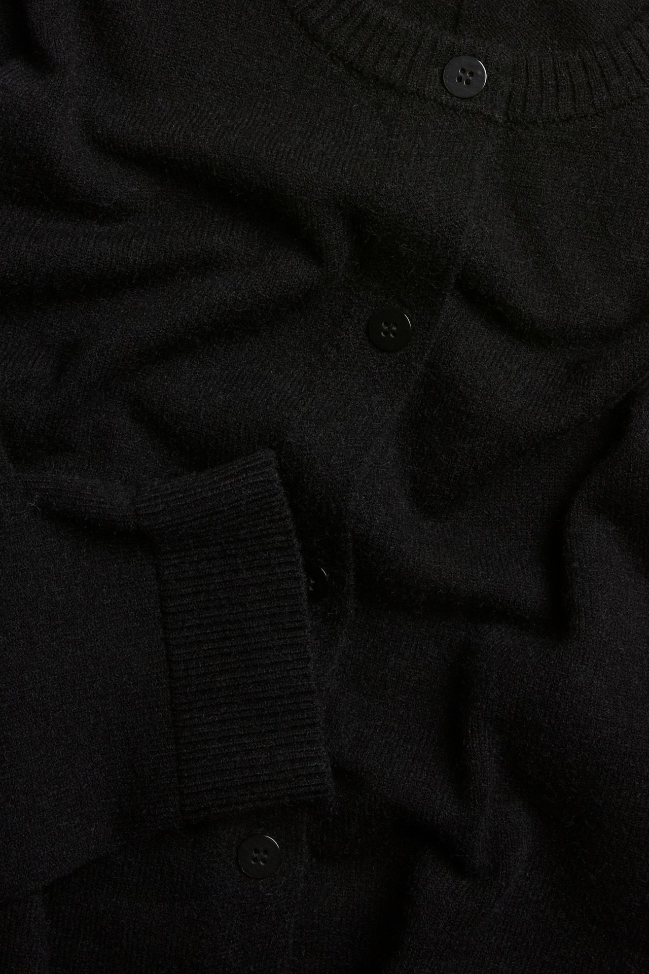 Zbliżenie na jednolity czarny dzianinowy kardigan z widocznymi czarnymi guzikami i prążkowanym mankietem.