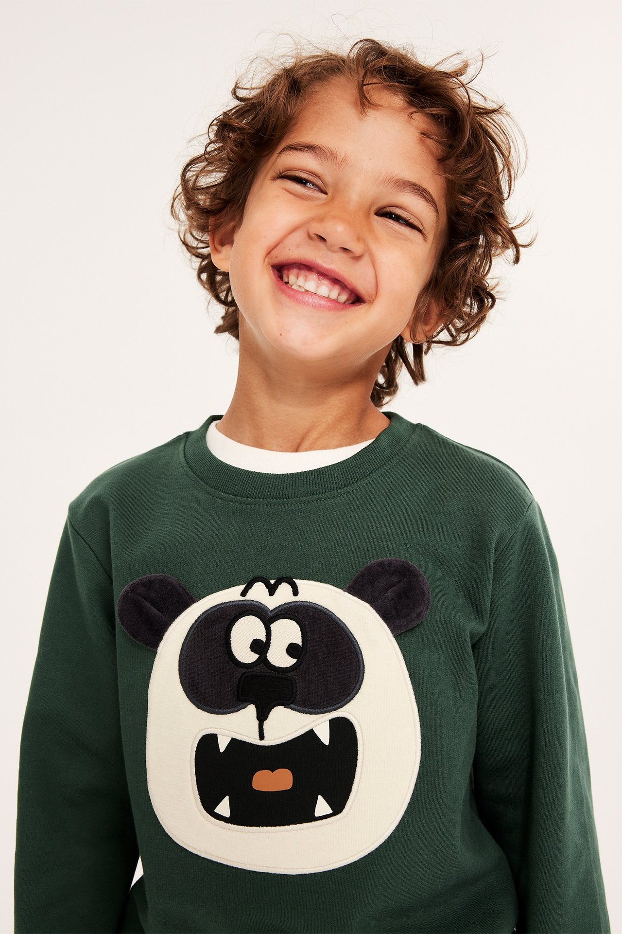 Widok z przodu: uśmiechnięte dziecko w zielonej bawełnianej bluzie z aplikacją pandy i uszami 3D.