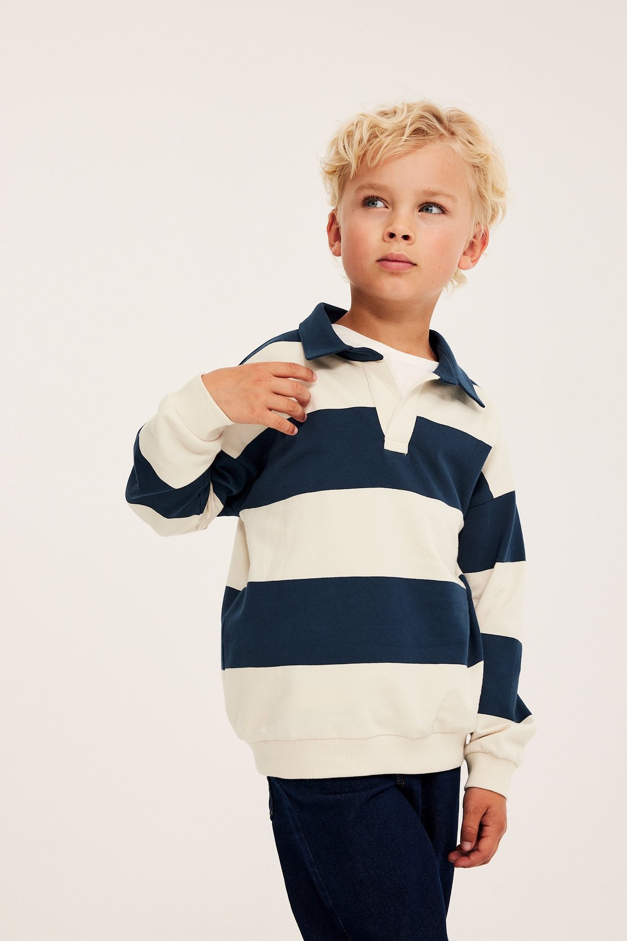 Framifrån: Ett barn bär en ljusbeige och mörkblå randig bomullssweatshirt med krage, vit t-shirt och mörkblå byxor.